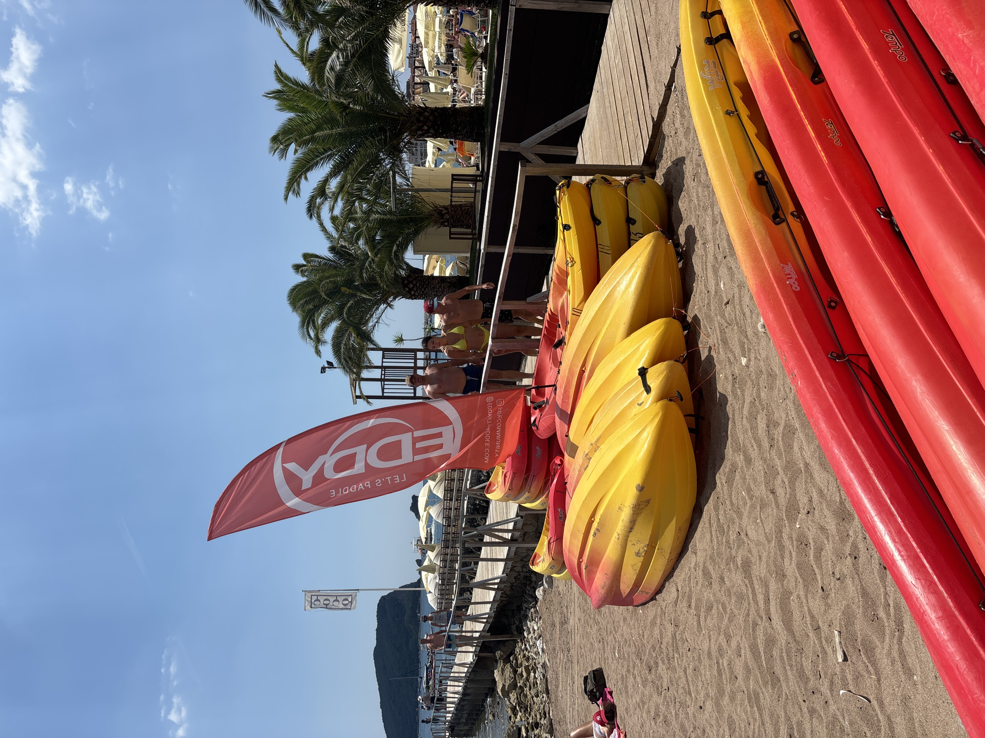 A group of kayaks, predominantly red and yellow, are neatly stacked on a sandy beach in Budva, Montenegro. The scene is set against a backdrop of lush palm trees and a clear blue sky with a few wispy clouds, suggesting a sunny day. A wooden boardwalk extends into the distance, lined with beach umbrellas and chairs, indicating a resort or recreational area.
Several people are visible on the boardwalk and near the water. Some are casually dressed, enjoying the beach atmosphere, while others are in swimwear. There's a sense of relaxation and leisure. A red flag with white lettering, partially unfurled, stands prominently in the foreground. The visible text on the flag appears to be "EDDY" and "LET'S PADDLE". A website address, "EDDA-LOVETE.COM", is also visible.
The time of day appears to be midday or early afternoon, given the bright sunlight and the shadows cast by the objects and people. The overall mood of the image is one of a pleasant summer day at a seaside destination, with opportunities for water activities.