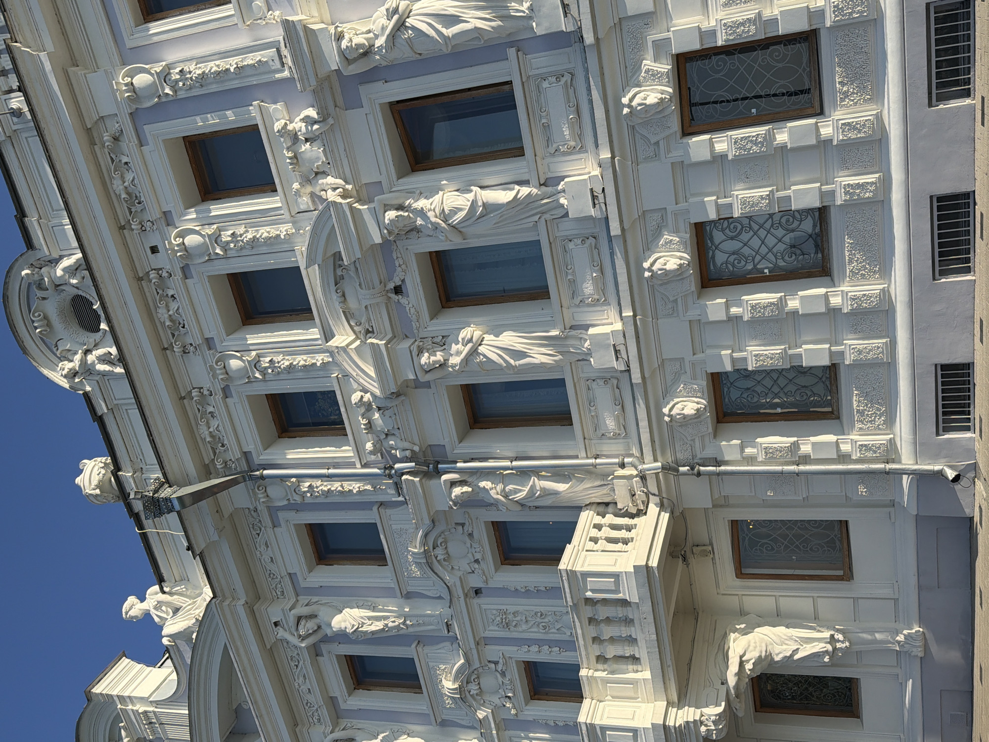 This is a low-angle, close-up photograph of the facade of a historic building in Nizhny Novgorod, Russia. The building exhibits ornate neoclassical architectural elements, characterized by extensive white stucco ornamentation against a pale lavender wall. Sculptural details, including statues of female figures in classical attire, putti, and decorative motifs such as garlands and lion heads, adorn the facade. Several windows with wooden frames and decorative wrought-iron grilles are visible. A section of a balcony with a balustrade extends from the upper floor. A silver downspout runs vertically along the facade. The clear blue sky serves as the background. The image captures the intricate details and grandeur of the building's exterior.