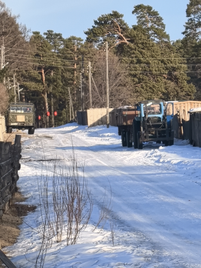 The image displays a snow-covered, unpaved rural road in Аблатуйский Бор, Russia, under a clear blue sky. On the left side of the road, an olive-green two-axle utility truck, possibly a GAZ-66, is stationary and oriented away from the camera. An individual is discernible in the driver's seat, and two circular red objects, likely taillights, are visible at the rear. On the right side of the road, a blue tractor, possibly an MTZ model, is also stationary and facing away from the camera, with its driver's side cab door open and an occupant visible within the cab. The tractor is towing a two-axle trailer with brown wooden side panels, which appears to be loaded with materials. The road surface consists of compacted snow and ice with visible vehicle tracks. The environment features tall coniferous pine trees and bare deciduous trees along both sides of the road, particularly in the background. Wooden utility poles with multiple power lines run parallel to the road. Various wooden fences or structures are situated along the roadside. In the immediate foreground on the left, several bare saplings emerge from the snow.