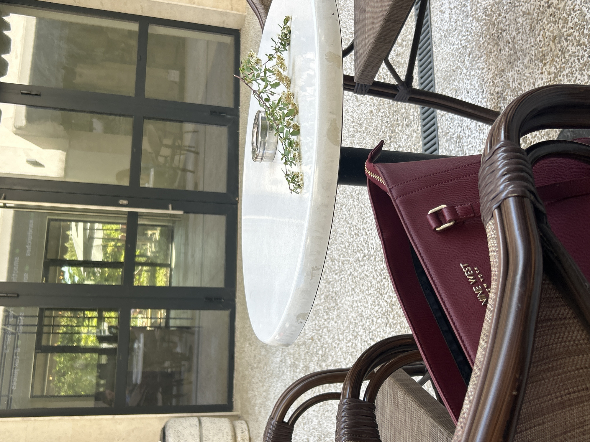 The scene depicts a café setting during daylight. A round, white table with a decorative sprig of flowers in a glass vase is the central element. A partially visible, red Nine West handbag rests on a brown, woven chair. Adjacent is a black-framed window, reflecting foliage and a glimpse of the interior. The location appears to be outdoors on a patio area with a textured, light-colored surface. The city is unknown.