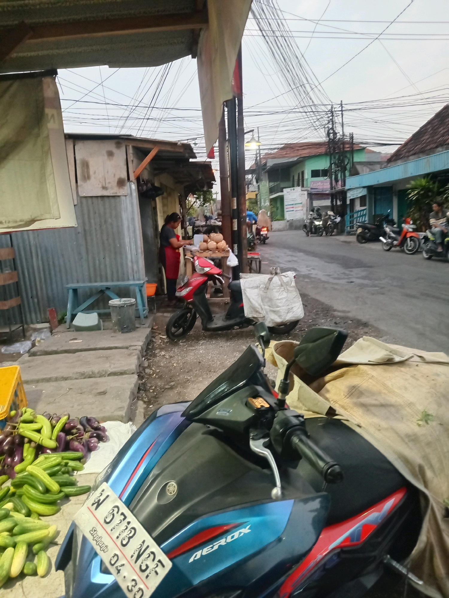 The image depicts a street scene in Kwangsan, Indonesia. In the foreground, a blue and red motorcycle is parked, partially covered with a tan cloth. The motorcycle has a license plate with the inscription "W 6713 NGE" and "Stephanie 04.30". To the left of the motorcycle, a pile of cucumbers and eggplants is displayed for sale on the ground. Behind the produce, a blue bench and a grey bucket are visible near a corrugated metal stall. A woman wearing an apron is standing inside the stall, interacting with a pile of round objects, possibly coconuts. Further down the street, other motorcycles are parked and people are seen walking and riding. The background features buildings with red tile roofs, overhead electrical wires, and a cloudy sky.