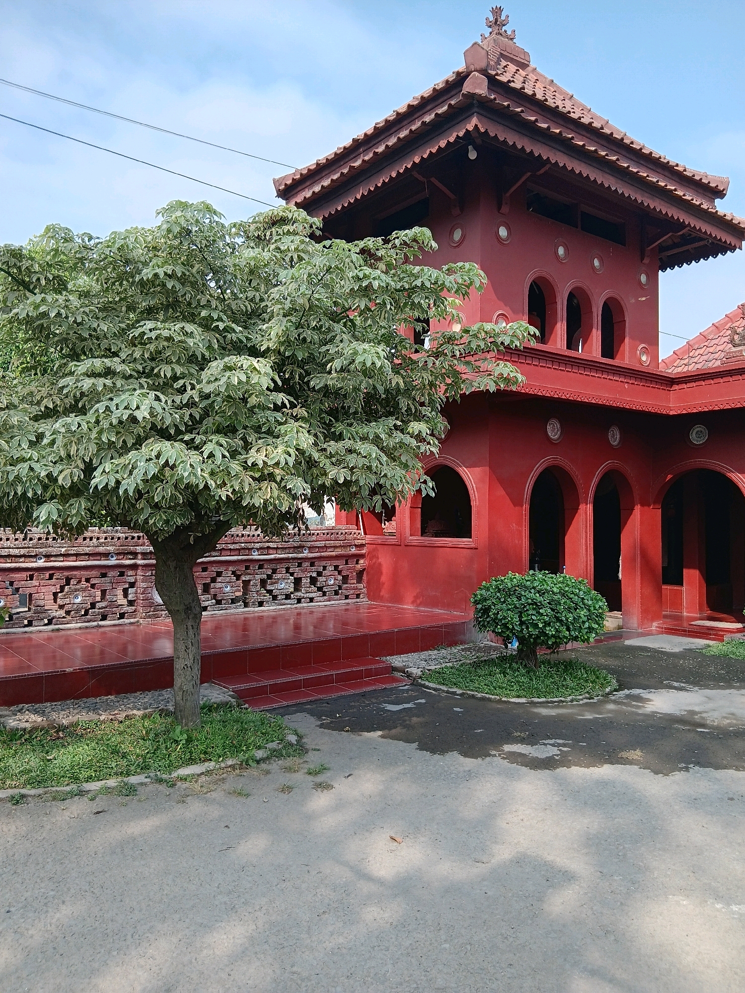 The image shows a view of the red building of the Sunyaragi Water Castle in Cirebon, Indonesia. The building is a historical site with distinct architectural features, including arched windows and a tiered roof. In the foreground, a tree with variegated leaves stands to the left, and a small, rounded bush is positioned in front of the building on the right. The ground is a mixture of paved surfaces and patches of grass, with some puddles of water reflecting the surroundings. The sky above is a clear blue with a few wispy clouds. The overall scene depicts the exterior of a historical landmark in Cirebon, Indonesia.