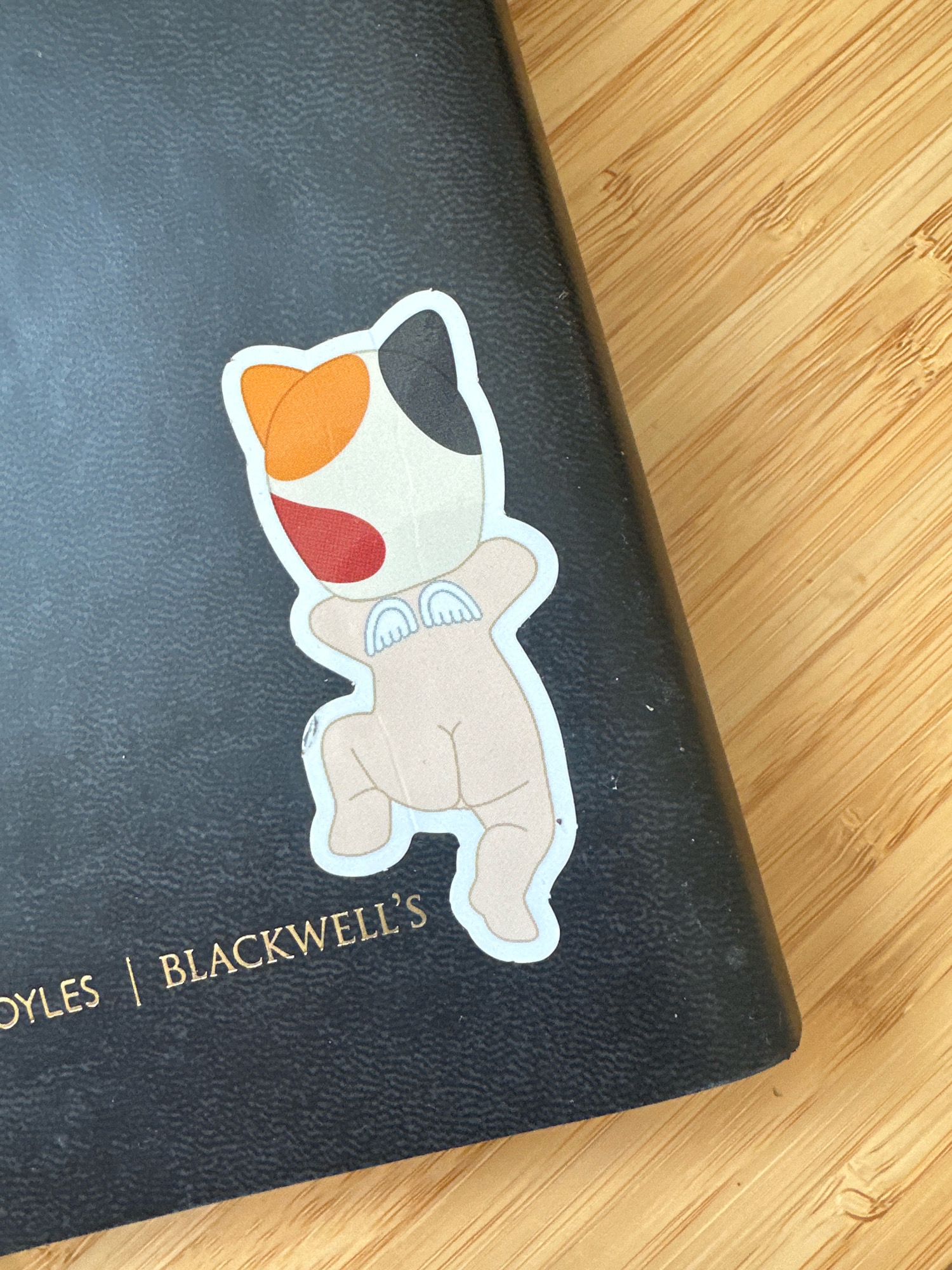 A calico cat sticker is placed on the cover of a black book with the embossed gold text "OYLES | BLACKWELL'S". The book is positioned on a light-colored wooden surface with a distinct grain pattern. The sticker depicts a stylized, anthropomorphic cat figure with orange, white, and black markings on its head, a red patch on its face, and small wing-like embellishments on its back. The cat figure appears to be in a playful or dancing pose, with its legs bent and arms outstretched. The wooden surface suggests an indoor setting, possibly a desk or table. There is no discernible indication of the city of Tunbridge Wells, United Kingdom.