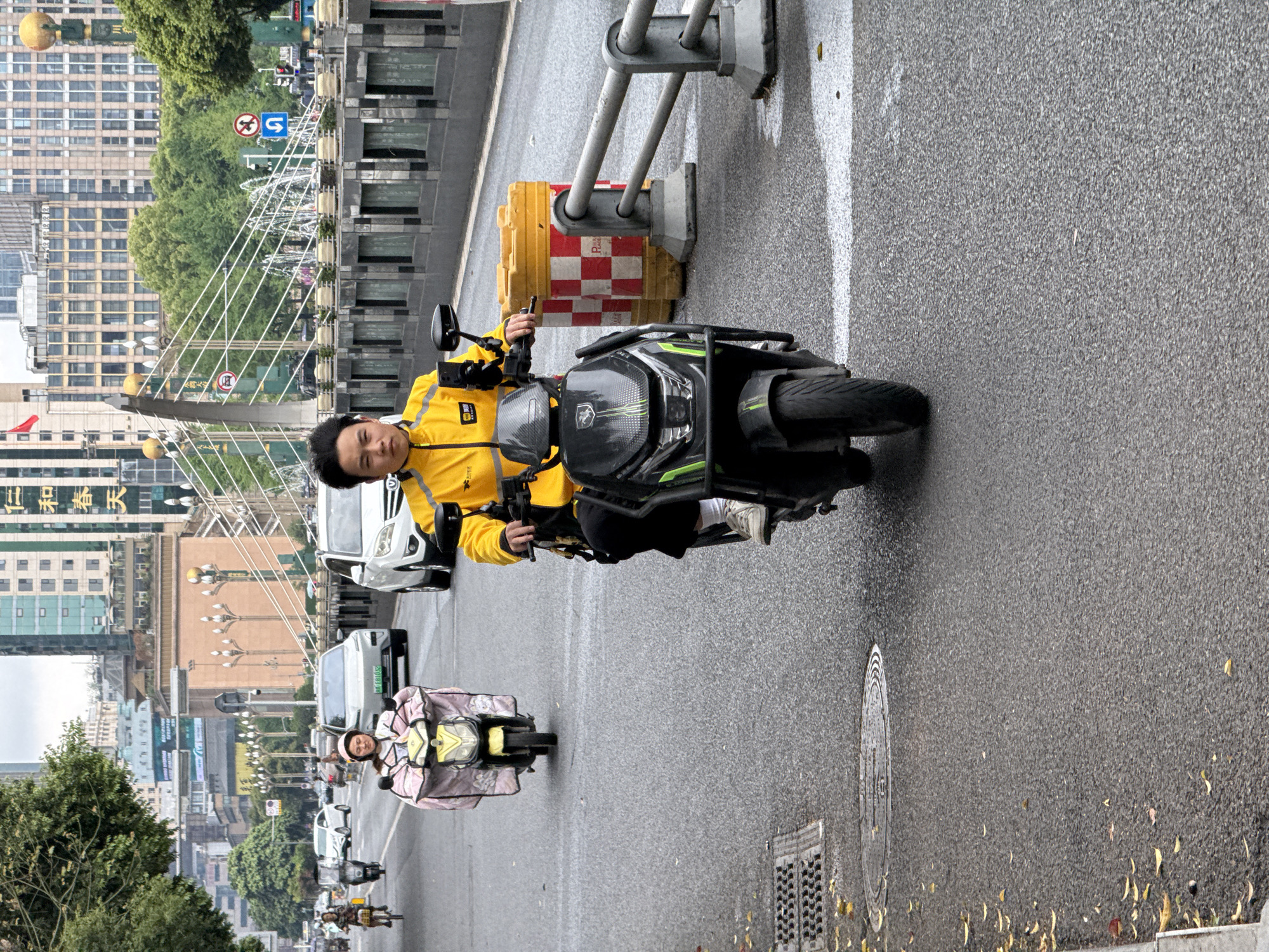 The image depicts a street scene in what appears to be the Qingyang District of a Chinese city, possibly Chengdu based on architectural cues. A man wearing a yellow jacket rides a dark-colored motorcycle directly towards the viewer. Behind him, another person on a smaller electric scooter with a pink protective cover is traveling in the same direction. Several vehicles, including cars and scooters, are visible further down the road, as well as pedestrians. Buildings are lining both sides of the street. A distinctive white cable-stayed bridge-like structure arches overhead. The street itself is wet, with some leaves scattered near the curb. Traffic barriers stand near the main motorcyclist on the right.