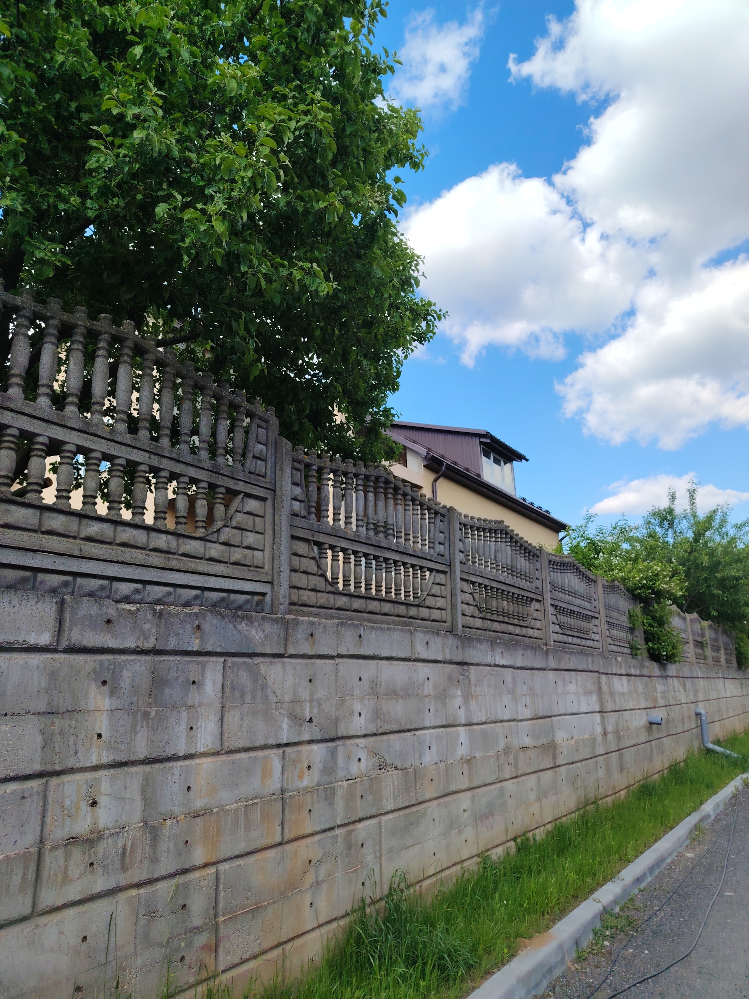 The image shows a concrete fence and wall bordering a street in the Бараўлянскі сельскі Савет region of Belarus. The wall is constructed from large, rectangular concrete blocks, with visible seams and some minor staining. Above the wall, a decorative concrete fence with a balustrade-like design is visible. A tree with lush green foliage partially obscures the fence on the left side of the frame. In the background, a house is partially visible, with a brown roof. The sky is blue with scattered white clouds. Grass grows along the base of the wall and along the edge of the sidewalk. A gray sidewalk borders the grassy area. The perspective suggests the photo was taken from a slightly low angle on the street.
