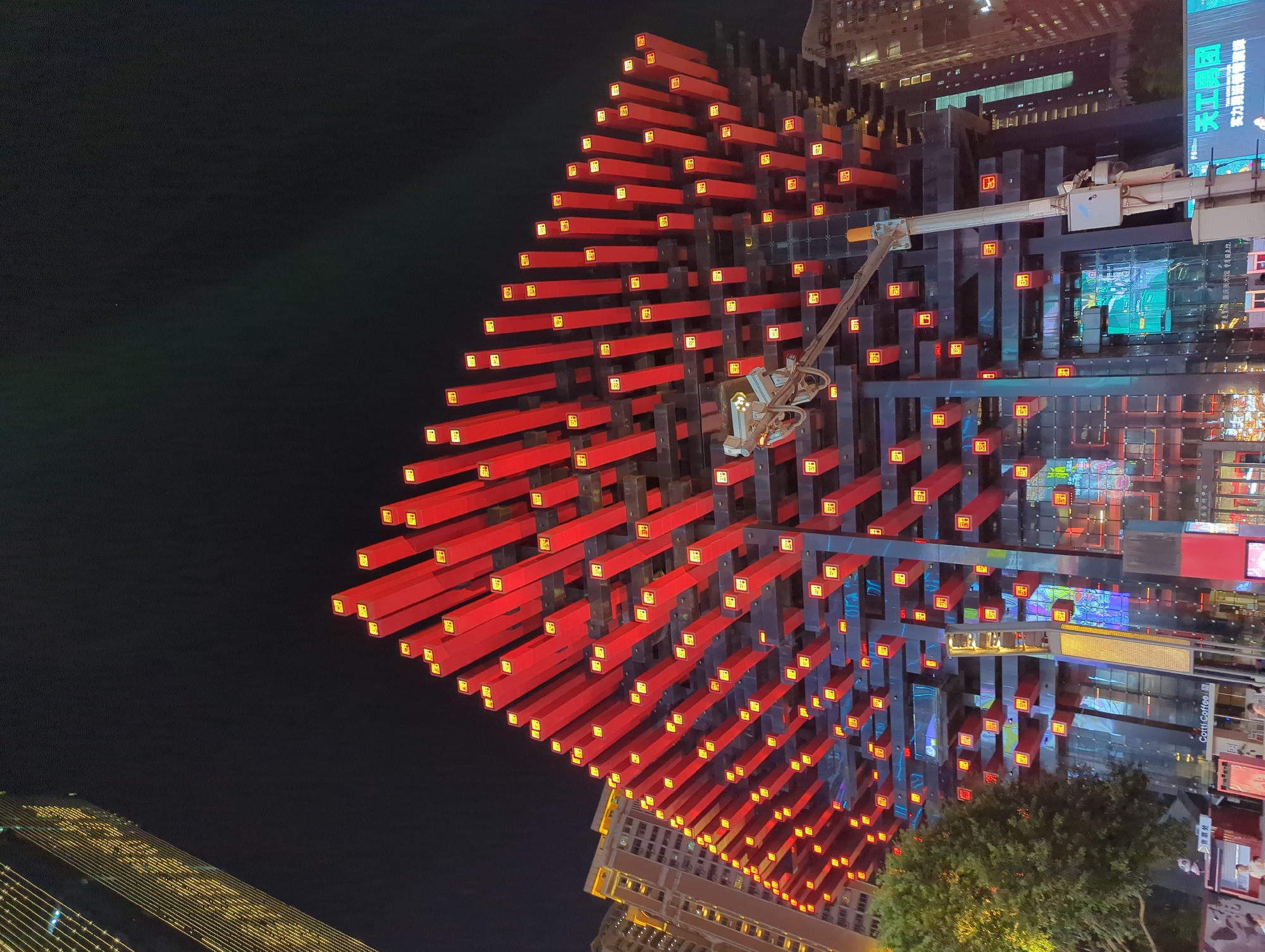 The image depicts the exterior of a modern building in Yuzhong District, China, at night. The building's facade is characterized by an intricate structure of red, rectangular beams, each illuminated by a small, square yellow light. These beams are arranged in a dynamic, staggered pattern, creating a visually striking effect. Below this illuminated structure, the building's lower levels appear to be clad in glass, reflecting the surrounding lights and the night sky. To the left of the main building, a tall skyscraper with a facade adorned with numerous yellow lights is partially visible, stretching upwards into the dark sky. A large, mature tree with dense green foliage stands to the left of the building, its leaves silhouetted against the illuminated structure. In the foreground, a traffic light pole with attached surveillance cameras is positioned in front of the building. Various signs and storefronts are visible at the base of the building, including one that reads "Cotti Coffee." Several indistinct figures can be seen near the entrance of the building, suggesting pedestrian activity. The overall scene is one of urban nighttime illumination, showcasing architectural innovation and a vibrant city environment.