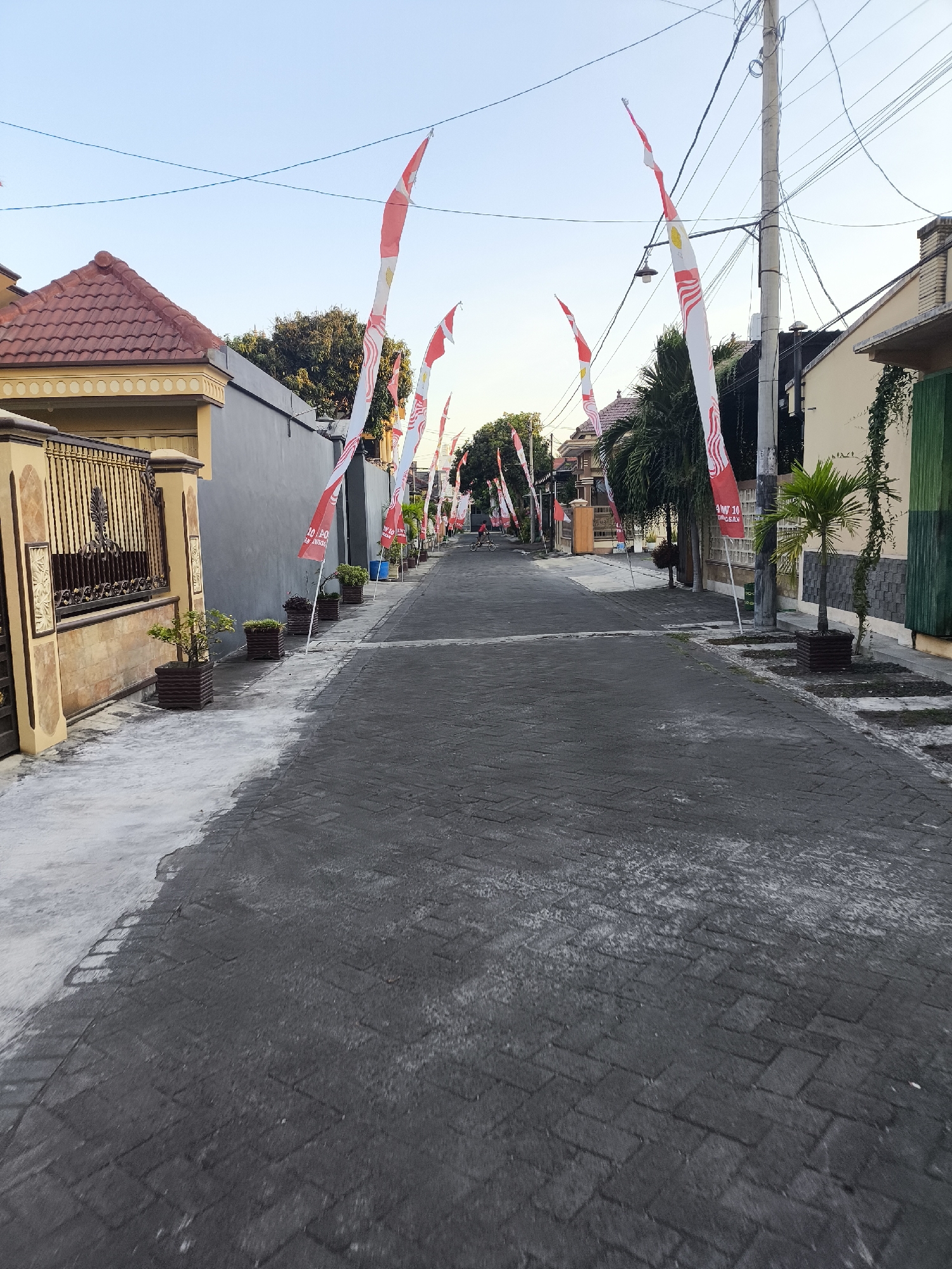 The image depicts a residential street in the city of Citrodiwangsan, Indonesia. The street is paved with dark rectangular bricks. Red and white Indonesian flags are prominently displayed on tall poles along both sides of the street, suggesting a national celebration or event. Houses with red tiled roofs and neutral-colored walls line the street. Planters with small green plants are situated along the sidewalk. In the distance, a person is visible riding a bicycle. The sky is clear and a pale blue, indicating it is either early morning or late afternoon. The overall scene is calm and orderly.