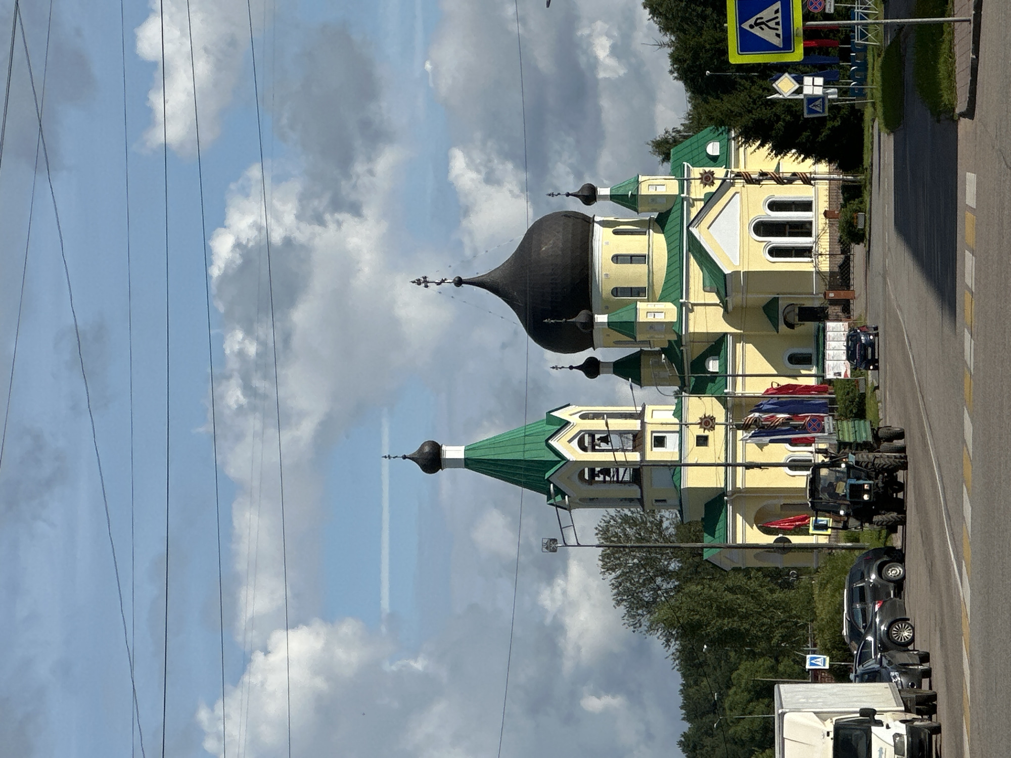 In the city of Slantsy, Russia, a yellow Orthodox church with green roofs and black domes stands prominently against a sky filled with white, puffy clouds. To the left of the church, a bell tower with a green spire and black dome is visible. A tractor towing a trailer is driving on a road in front of the church, with several cars parked nearby. A white truck is also on the left side of the road. Traffic signs, including a pedestrian crossing sign, are visible on the right side of the road. The surrounding area is lined with trees, and power lines stretch across the sky.