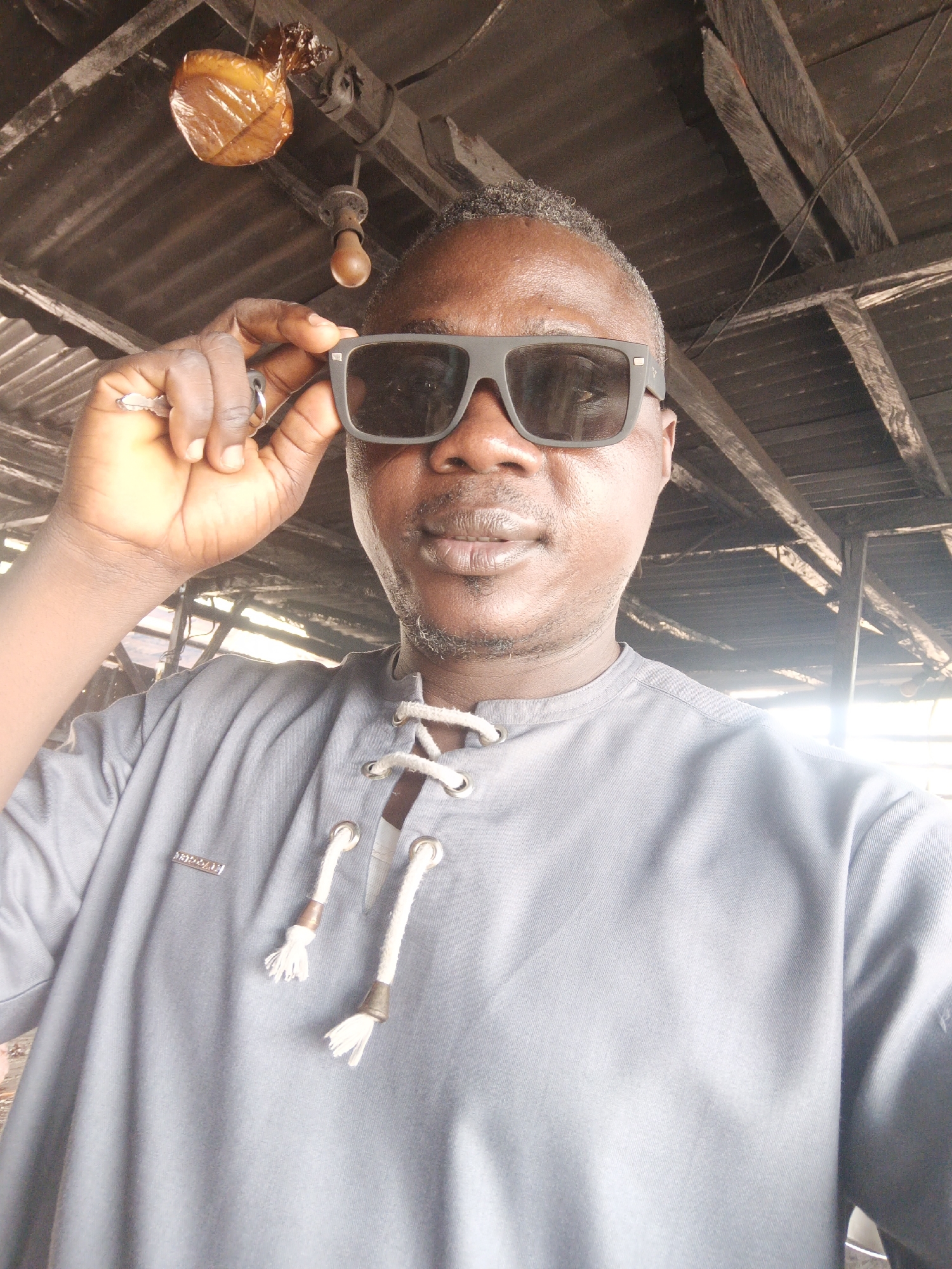 A man wearing sunglasses and a gray shirt is posing for a selfie in what appears to be an indoor, possibly commercial, location in Akwanga, Nigeria. The man is holding the sunglasses with his left hand, which also has rings on its fingers. In the background, there is a rustic roof structure, an obscured object in a plastic bag, and a hanging lightbulb. A logo is visible on the man's shirt. The lighting is somewhat dim.