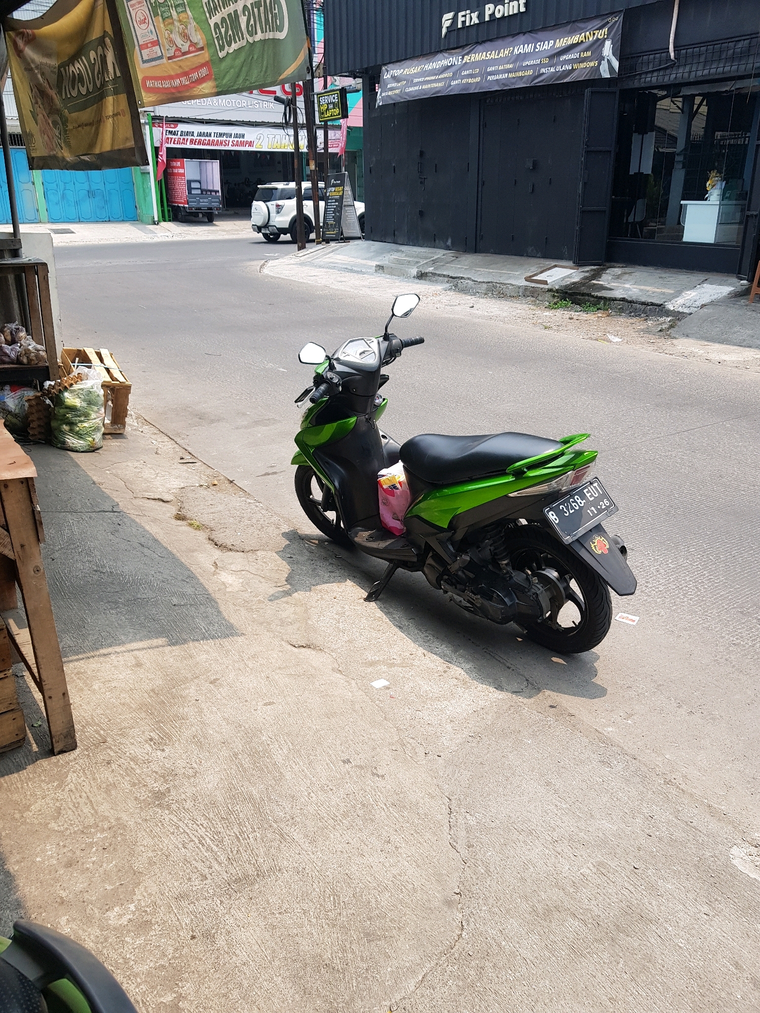 The image depicts a street scene in Bekasi, Indonesia. A green and black motorcycle is parked on the right side of the road. The motorcycle has a license plate that reads "B 3268 EUT 11-26". In the background, there is a black building with a sign that says "Fix Point". To the left of the building, there is a white SUV parked and a blue building with rolled-up shutters. In the foreground, there is a concrete sidewalk with some debris. The overall lighting suggests it is daytime.
