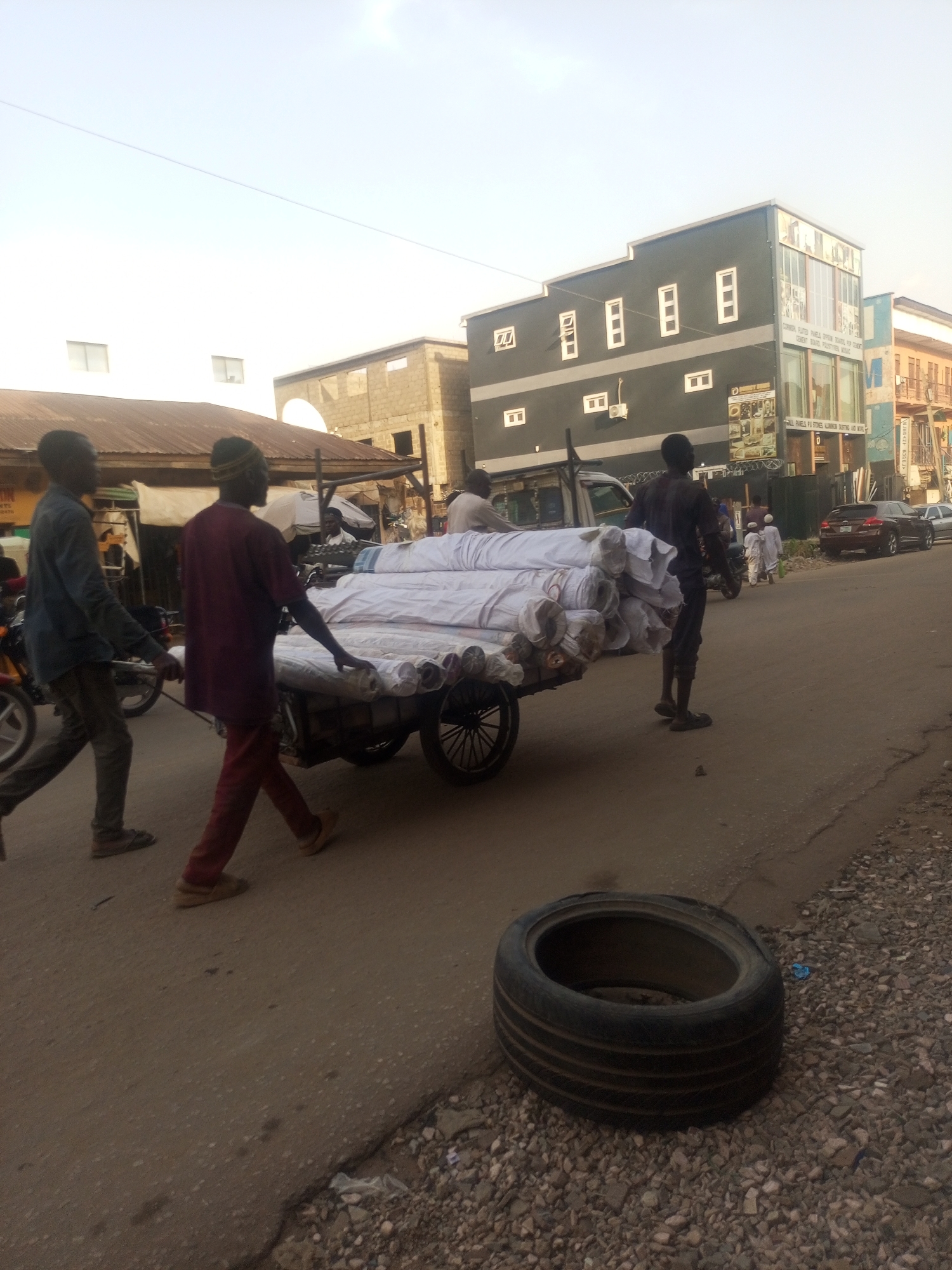 Three men are pulling a wheeled cart loaded with numerous rolls of white fabric down a paved road. The scene appears to be an urban street in Maiburiji, Nigeria. The men are dressed in casual clothing. One man pulls the cart from the front, another is walking along the cart, and the third man is walking behind it. A discarded tire lies on the right side of the road. Various buildings can be seen in the background, including a grey two-story building and other commercial structures. Motorcycles and cars are visible on the street as well. Other people, including two men in white robes, are walking on the opposite side of the road.