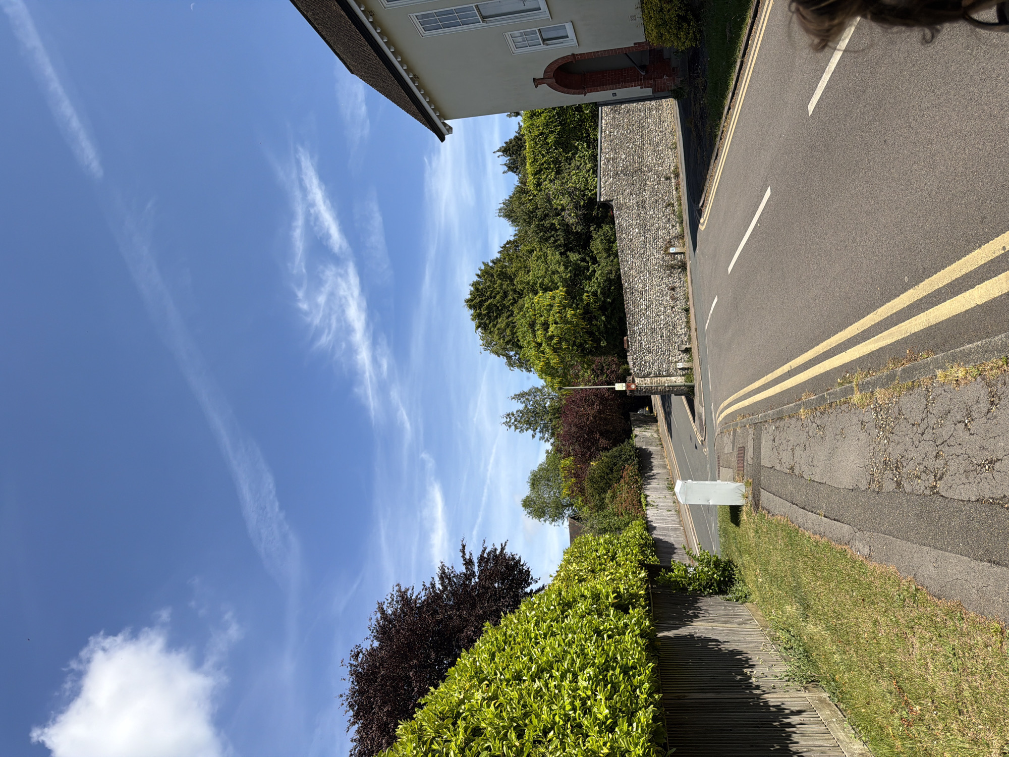 The image depicts a street scene in Guildford, United Kingdom, taken during the daytime with clear blue skies and scattered cirrus clouds. The perspective is from an elevated position, possibly from a window or balcony. A two-story building with a red brick archway over the entrance is visible at the top. A stone wall runs along the right side of the frame adjacent to a paved road with double yellow lines. A green hedge lines the lower portion of the frame, with some red foliage visible amongst it. The road is cracked and worn, and there's some grass and weeds growing along the curb. A white, rectangular structure, possibly a utility box, sits on the edge of the road. In the background, a variety of trees and foliage are visible. A person's head is visible in the top right corner of the image.