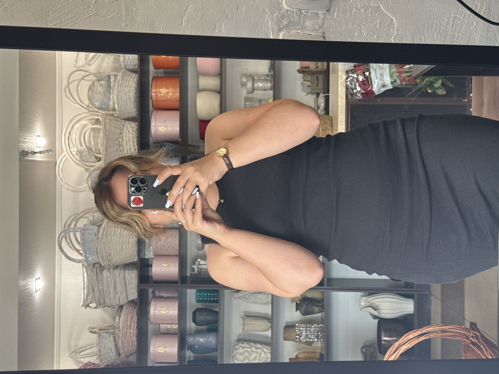 The image is a mirror selfie taken by a person wearing a black ribbed dress and a wristwatch. The person is holding a smartphone in their left hand. The background, reflected in the mirror, shows shelves filled with various items, including woven baskets, cylindrical containers in shades of orange, pink, and cream, and decorative vases. There are also red roses wrapped in plastic visible on a shelf in the upper right of the reflection. The setting appears to be a retail environment, possibly a gift shop or a boutique, given the display of merchandise. The lighting suggests an indoor setting, likely during daytime. There is no discernible text in the image.