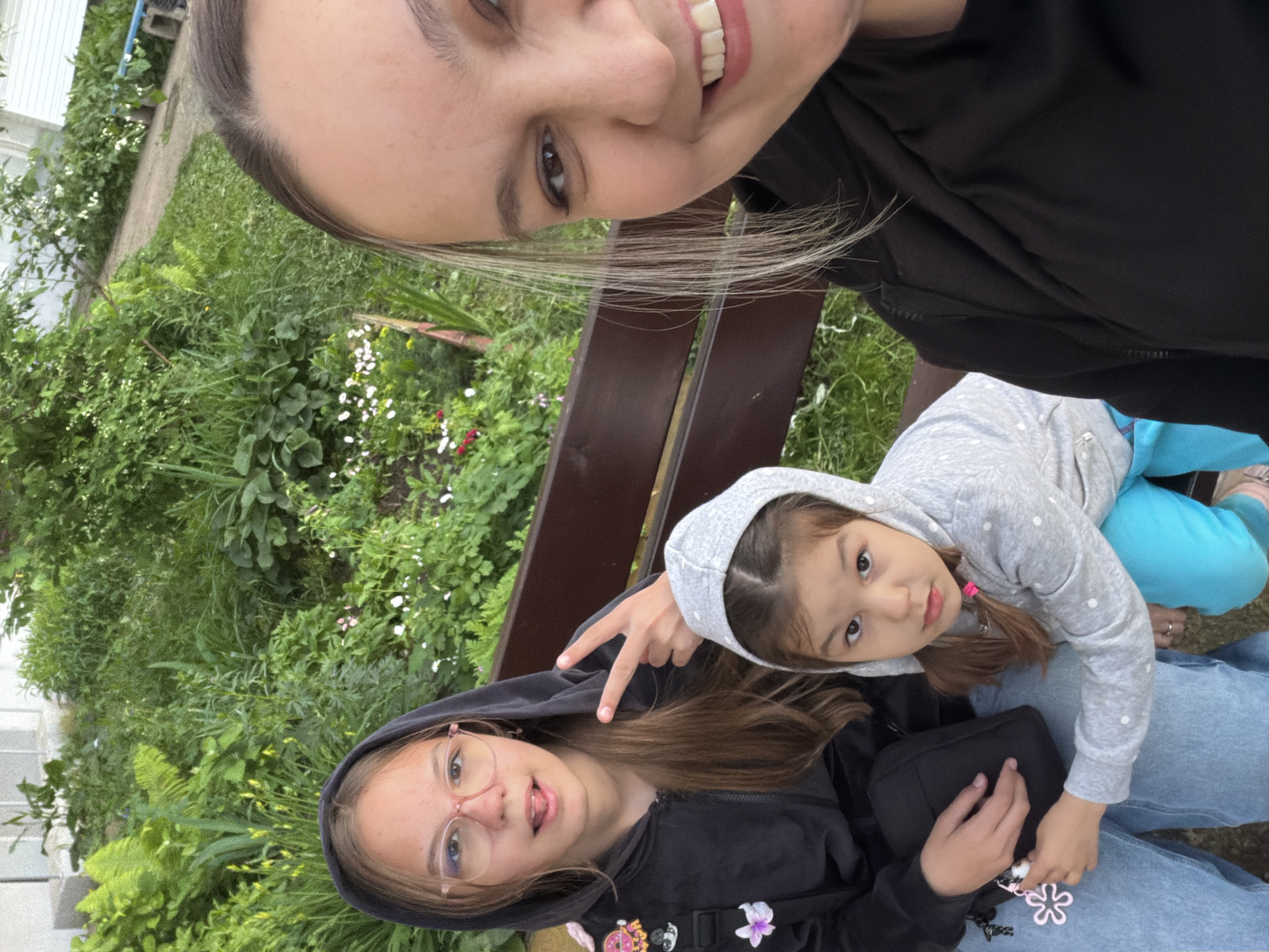 This outdoor selfie captures three females across different generations, likely a family, in what appears to be a garden or park setting. The image is taken from a slightly elevated perspective, with the person holding the camera, an adult or young adult, positioned at the top of the frame. She has light-streaked hair and is smiling broadly at the camera.
Below her, a teenager sits on a dark brown wooden bench. She is wearing a black hooded sweatshirt, light blue jeans, and light pink-rimmed glasses. With a playful expression, she is seen making a "peace" sign with her right hand and slightly sticking out her tongue. A black shoulder bag adorned with several colorful pins and charms, including a pink flower and a black-and-white yin-yang symbol, is visible on her side.
Leaning into the teenager's lap is a young child, dressed in a grey hooded sweatshirt with white polka dots and bright blue pants. The child has brown hair, partially tied with a pink elastic, and looks directly at the camera with a calm, curious gaze.
The background is abundant with lush green foliage, including various plants, bushes, and grass, interspersed with small white and red flowers. A building with white siding can be partially seen on the left side of the frame. The lighting suggests it is daytime, possibly overcast or late afternoon, creating a soft, even illumination. The scene conveys a relaxed and cheerful atmosphere, capturing a moment outdoors in Chaikovsky, Russia. There is no visible text in the image.