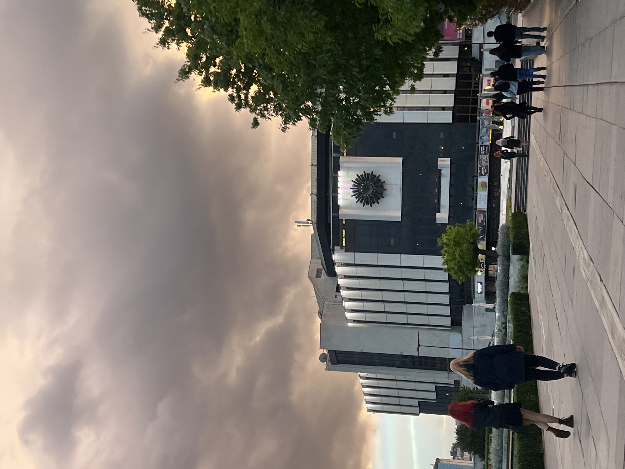 The image captures a view of the National Palace of Culture in Sofia, Bulgaria, under a sky filled with dark, stormy clouds. The building's distinctive architecture, featuring alternating bands of dark and light-colored panels, is prominent. A large sun-shaped emblem adorns the center of the darker section.
In the foreground, a paved walkway is visible, and a group of people are walking away from the viewer and towards the building. Closer to the viewer, two women are walking in the opposite direction. The woman closest has red hair, wears a black dress and brown shoes. The other woman has long blonde hair and is wearing black leggings and a black sweater. A line of trees is planted along the edge of the walkway.