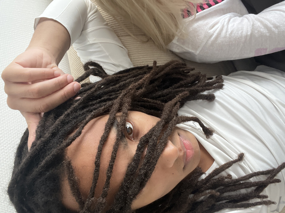 This indoor image, located in Paris, France, features two individuals. The primary subject, positioned on the left, has dark skin and dark brown dreadlocks, several strands of which extend across and partially obscure their face. Their left eye, which is brown and clearly visible, is directed towards the viewer. The subject's left hand, with fingers slightly spread, rests near the top of their head amidst their dreadlocks. They are attired in a white, long-sleeved top with a draped neckline. To the right, a second individual is partially visible, identifiable by blonde hair and a portion of their upper body clad in a white long-sleeved shirt. A pattern of pink and black is discernible beneath or integrated into this white garment. Both individuals appear to be seated, with a light-colored, vertically ribbed upholstered surface, consistent with a sofa or padded chair, forming the background. The scene is evenly illuminated.