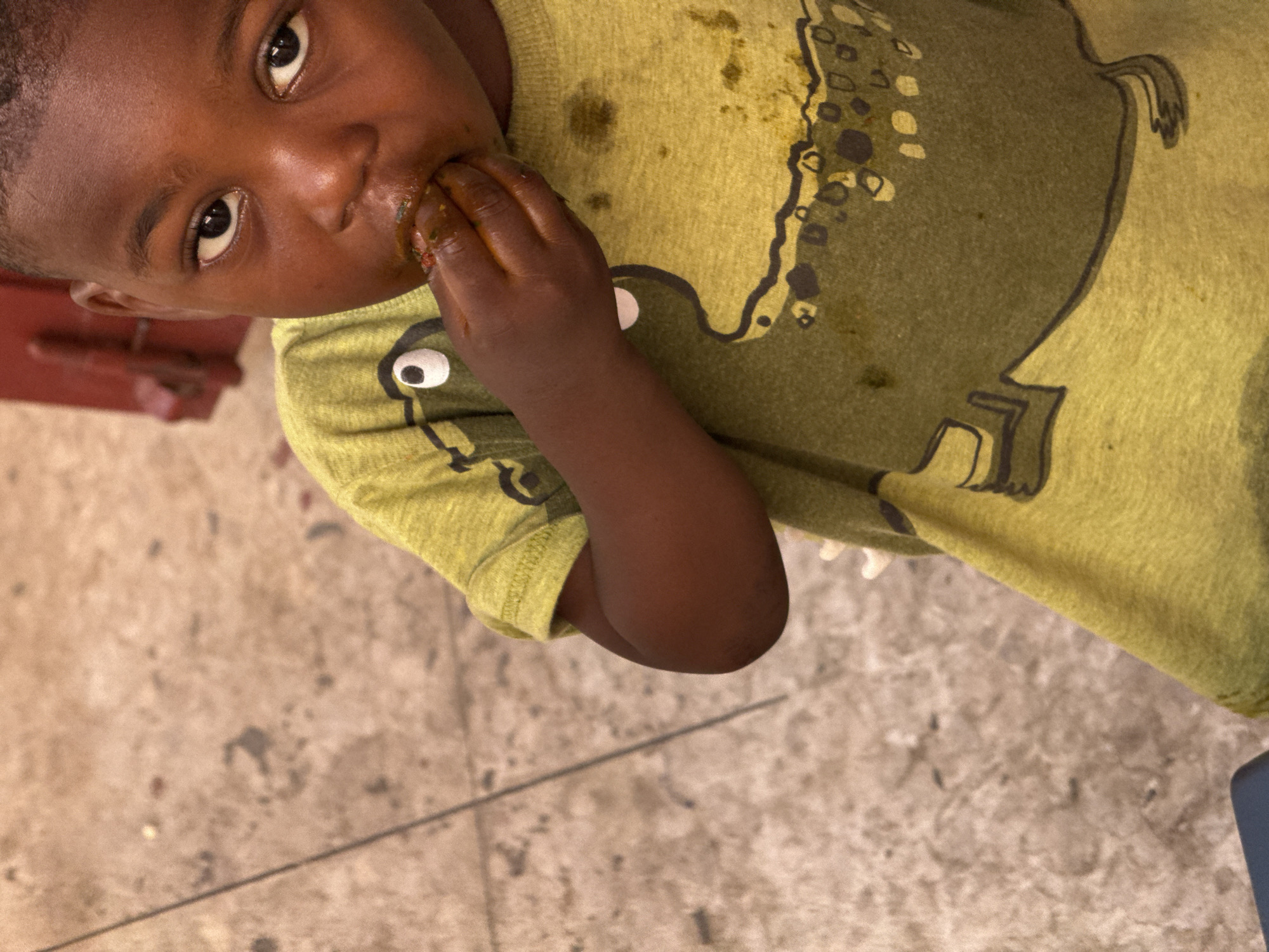 A young boy with dark skin is eating food with his hands. He is wearing a light green t-shirt with a dark green dinosaur graphic. The t-shirt has some food stains on it. The background is a concrete floor with some debris. Part of a red object is visible in the upper left corner. The location is likely Abuja, Nigeria.