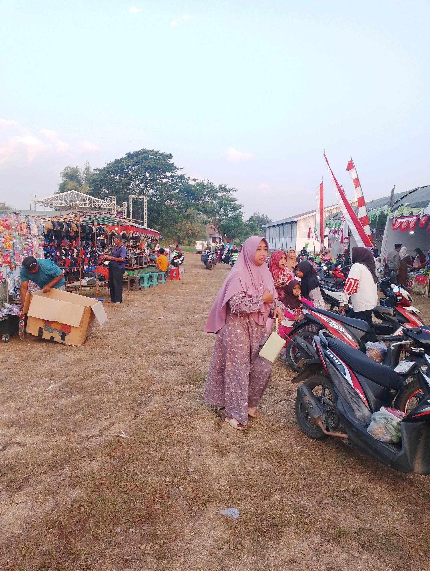 An outdoor market scene in Joho, Indonesia, features a variety of stalls and a dirt ground. In the foreground, a woman in a lavender hijab and patterned dress walks, carrying a light-colored bag. Several motorcycles are parked to her right. Further into the scene, a man in a teal shirt is handling a cardboard box, and another man in a blue shirt and dark pants stands near a stall displaying caps and accessories. In the background, tents and structures suggest further market activities, with Indonesian flags visible, and trees and buildings framing the scene under a bright sky.