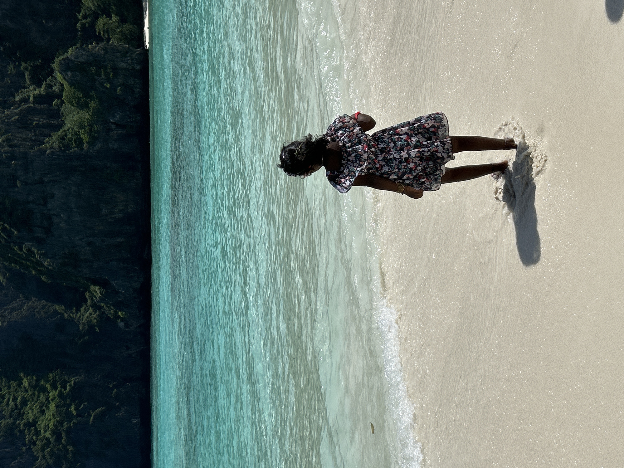 A young child wearing a floral dress stands on a sandy beach, with the turquoise waters of the Andaman Sea lapping at their feet. The child is facing away from the camera, looking out at the sea. In the background, a dramatic limestone cliff covered in lush greenery rises from the water. The scene appears to be a tropical paradise, likely in Ao Nang, Thailand, known for its stunning beaches and karst landscapes. The sun is bright, casting a shadow of the child on the sand. A few small waves are breaking on the shore, creating a white foam.
