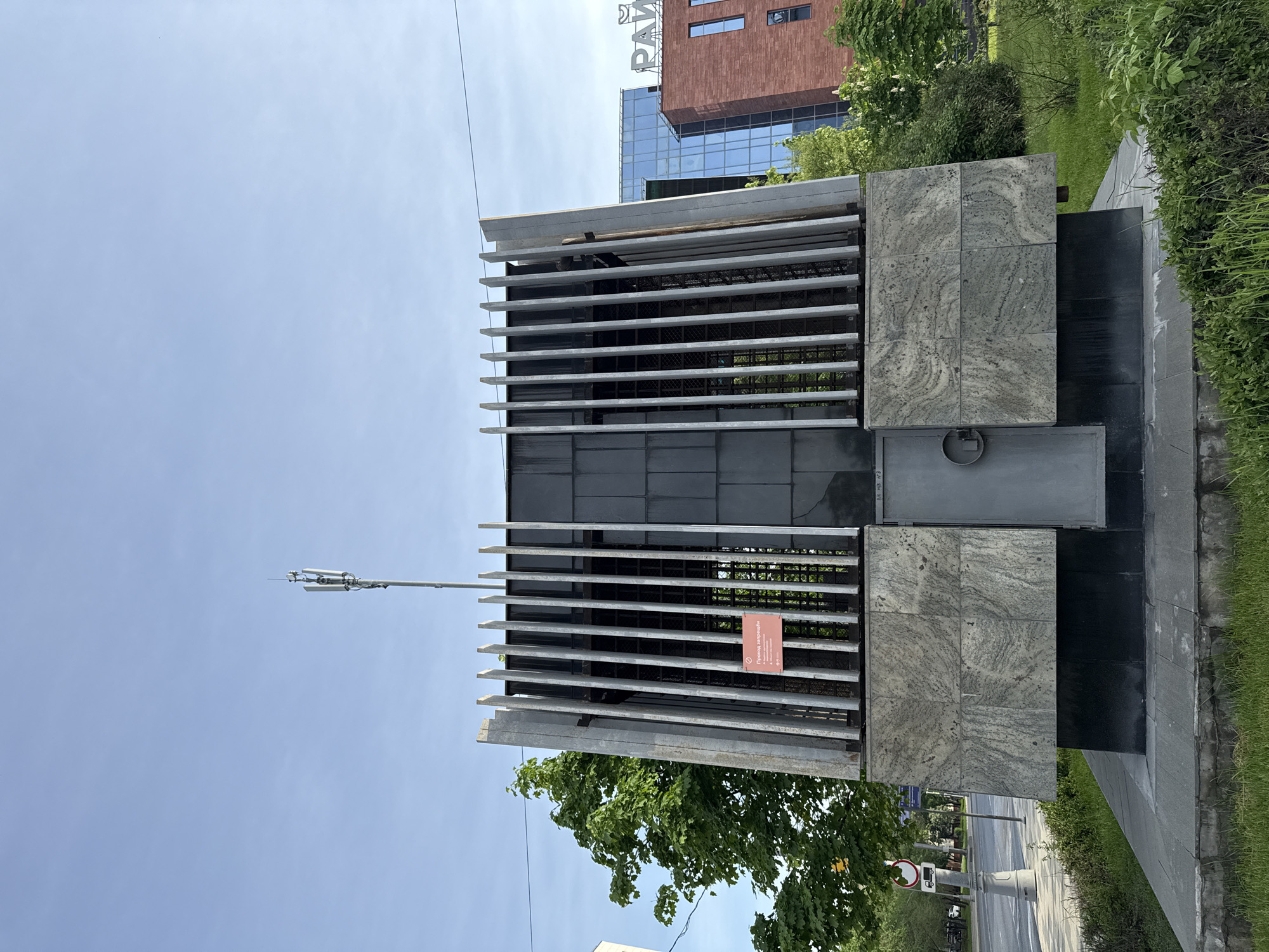 The image shows a modern architectural structure in Moscow, Russia. The structure is constructed of gray stone and metal, with a grated metal section covered by long metal bars. A metal door is seen on the right side of the structure. A sign written in Russian is visible within the grating. Atop the structure, a communications antenna is installed. In the background, a building with the sign "PAVILION" is partially visible. Street signs and trees are visible on the right side of the image, with a blue sky above. No people are visible in the image.