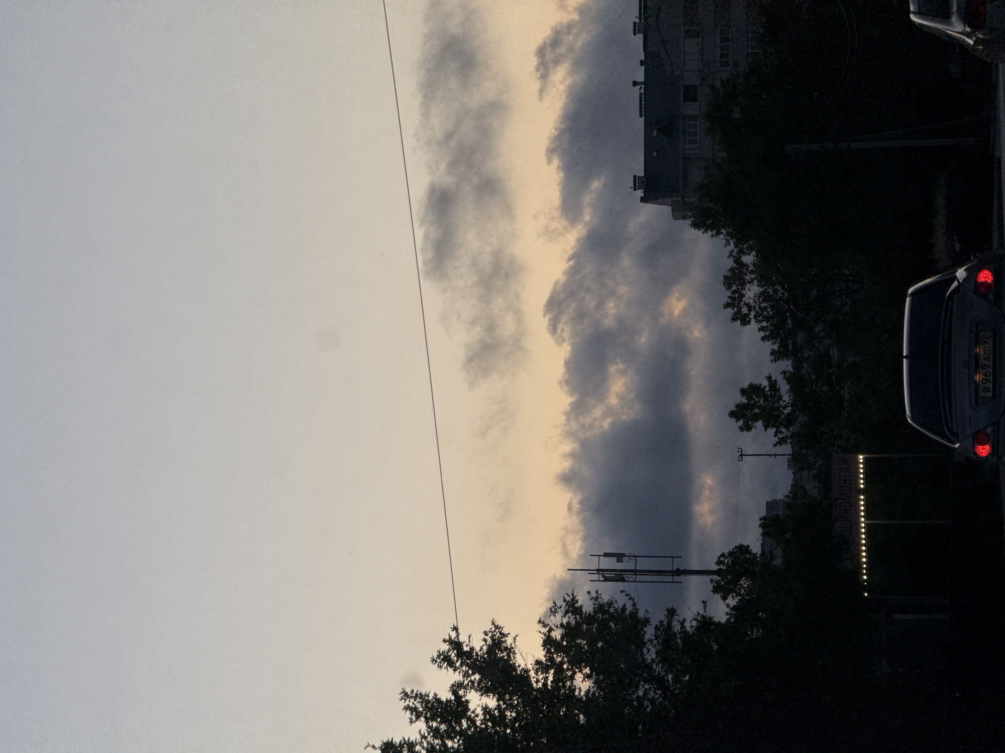 The image is an outdoor, slightly angled, eye-level shot of a city scene at dusk. The sky is a gradient of pale blue to pale orange with scattered dark clouds. A thin, dark power line bisects the upper portion of the frame horizontally. In the mid-ground, silhouetted against the sky, is a large tree on the left, a cell tower, and a multi-story building on the right. Further down, more trees and buildings are visible, also silhouetted. In the foreground, two cars are visible. The car on the left is a silver sedan with its red taillight illuminated, parked on a road. The car on the right is partially visible and appears to be a dark sedan. In the lower-middle ground, a sign with lights is visible, with Cyrillic text that appears to read "ШИНОМОНТАЖ", indicating a tire fitting service. The overall atmosphere is subdued and quiet.