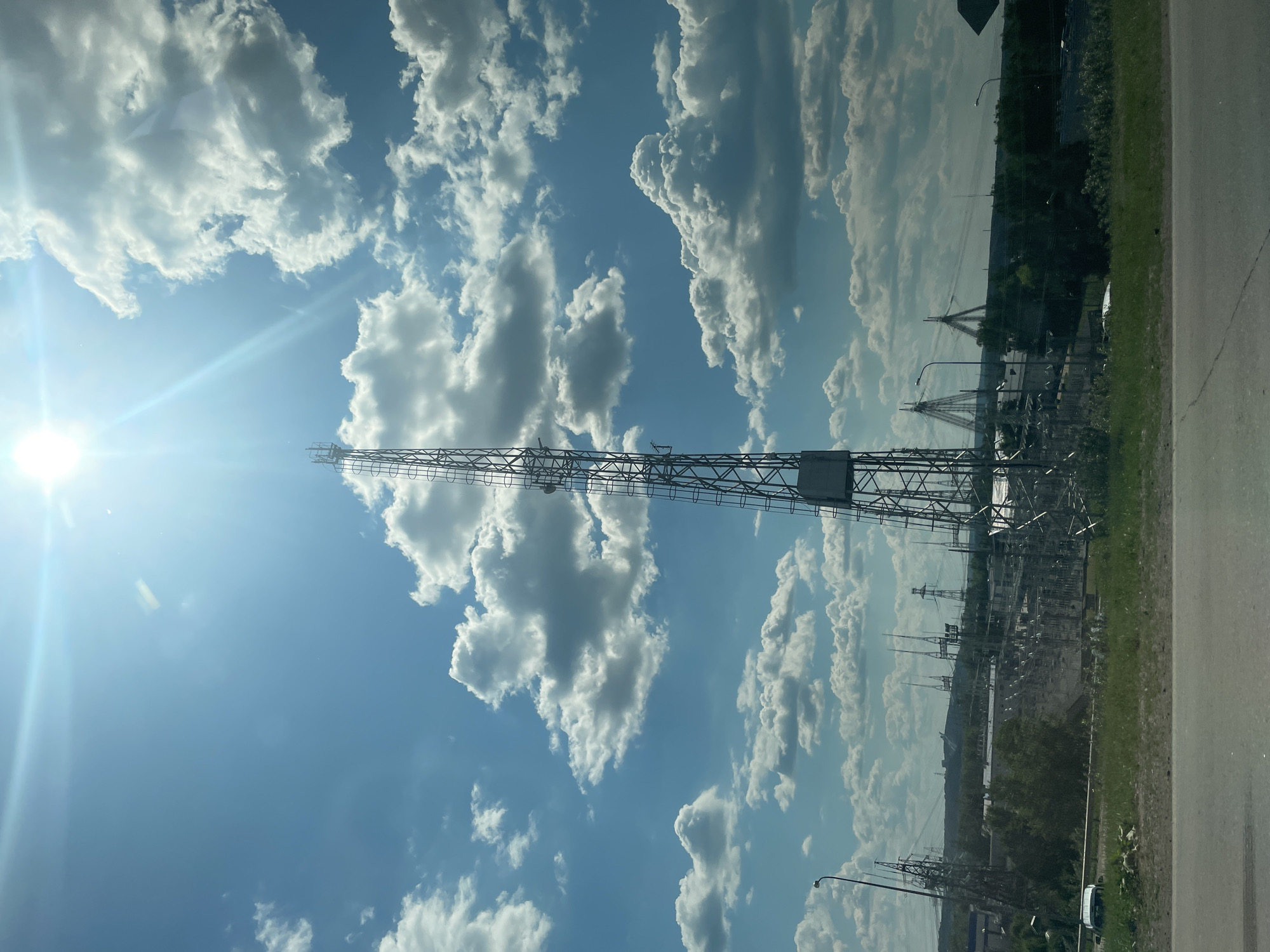 A tall, lattice communications tower stands prominently against a bright blue sky dotted with fluffy white clouds. The sun, high in the sky, creates a lens flare effect. In the background, a power substation with numerous electrical towers and lines stretches across the landscape. The area appears to be industrial or semi-industrial. The foreground shows a grassy verge and a paved surface, suggesting a roadside view. The location is identified as Chaikovsky, Russia, an industrial city known for its power generation facilities and located on the Kama River. There are no people visible in the image.