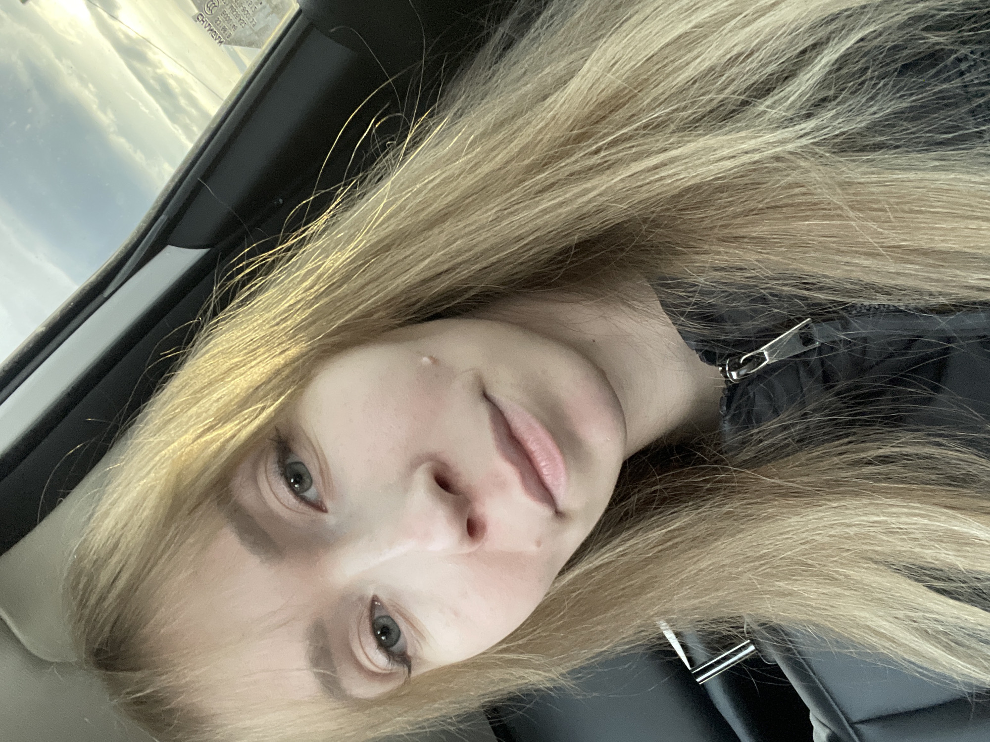 A close-up, angled portrait features a young woman with long, straight, light blonde hair. Her face is visible from the chin up, and she looks directly at the camera with light-colored eyes. Her expression is neutral. A small blemish or mole is visible on her left cheek. She is wearing a dark, possibly black, zippered jacket, with a silver zipper pull visible near her neck and shoulder.
The setting appears to be inside a vehicle, likely a car, indicated by the car seat or headrest visible behind her, and a car window frame in the upper left. Through the car window, an overcast or partly cloudy sky is visible, suggesting it is daytime. On the window, reversed and partially obscured text and numbers are faintly visible, resembling manufacturer's markings or a sticker (e.g., "CMVOHV," "0000"). The photo is taken at an upward angle, likely a selfie.