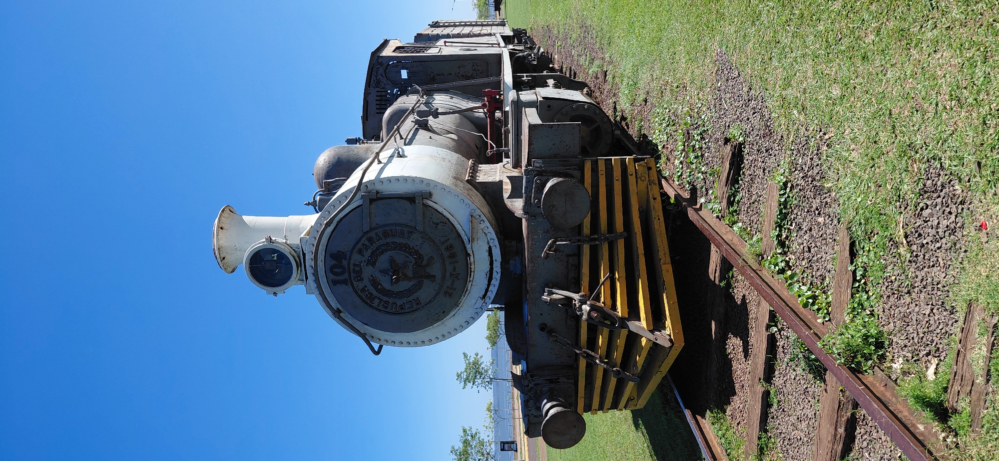 This is a full-length, eye-level, vertical shot of a vintage steam locomotive, painted primarily in white and grey, parked on a section of train tracks. The locomotive is positioned at an angle, facing slightly to the right, with its front prominently displayed. A bright blue sky with no clouds serves as the backdrop. The train's circular front emblem is visible, displaying the number "104" and text that reads "REPUBLICA DEL PARAGUAY" and "21-X-1961." The front of the locomotive features a yellow and black striped buffer beam. The train is situated on tracks with weathered wooden sleepers and a gravel ballast. To the left of the tracks, there is a patch of green grass, and beyond that, a body of water (likely the Paraná River) and trees lining the shore. In the distance, a few more trees and a clear blue sky are visible. The scene appears to be located in Encarnacion, Paraguay, as suggested by the text on the locomotive. There are no people or animals visible in the image. The overall impression is one of a historic artifact preserved in an outdoor setting.