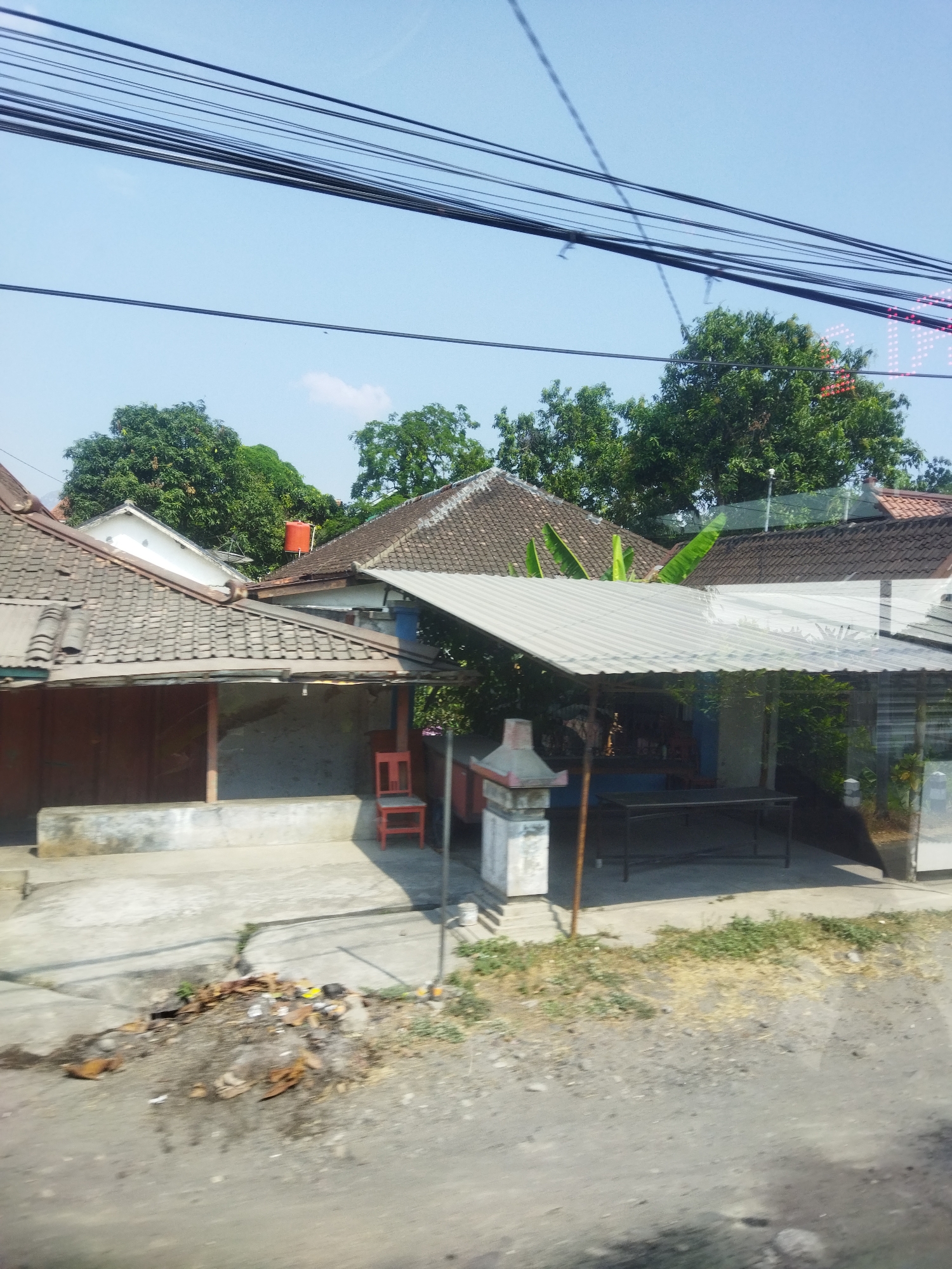 In Gemantar, Indonesia, a view captured from ground level shows a residential area with several houses. The foreground is dominated by a dusty, unpaved road with debris scattered on its surface, including leaves and small pieces of trash. Behind the road, a concrete pathway leads to a series of buildings.

The structures exhibit a variety of roofing materials, primarily gray tiles. One house in the left midground has a weathered, tiled roof and a brown wooden door. Adjacent to it, a smaller, open-sided structure is supported by a single pole and covered with a corrugated metal roof. A bright red chair is placed outside this structure. Further back, more houses with tiled roofs are visible, interspersed with lush green trees. In the upper background, a bright blue sky is crisscrossed by numerous overhead wires. A prominent orange water tank sits atop one of the houses. In the far right background, a sign with bright red lights is partially visible, suggesting a commercial establishment. The scene is illuminated by bright sunlight, casting shadows on the ground.