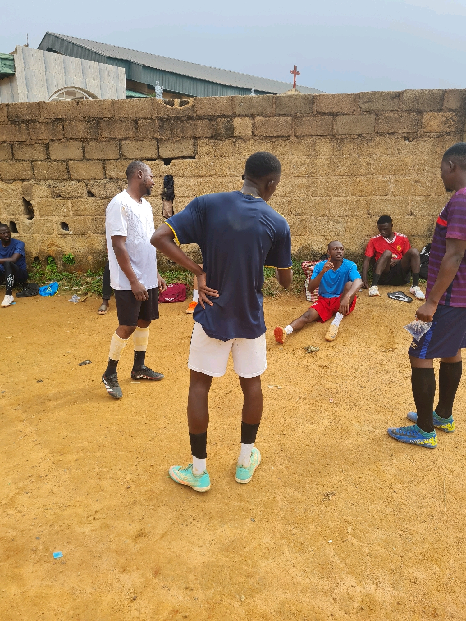 A group of young men are gathered in an outdoor area with a dirt ground. They appear to be in a casual setting, possibly after a sports activity, as some are wearing athletic attire and soccer cleats. One man in a blue t-shirt and red shorts is sitting on the ground and appears to be speaking, gesturing with his right hand. Another man in a white t-shirt and black shorts is standing and facing away from the camera, with his left hand on his hip. Other individuals are also visible, some standing and some sitting, in various athletic wear. The background features a brick wall, a building with a metal roof, and a cloudy sky. The overall scene suggests a relaxed gathering of individuals outdoors. The context of Jos, Nigeria, as the location implies the setting is likely within this city.