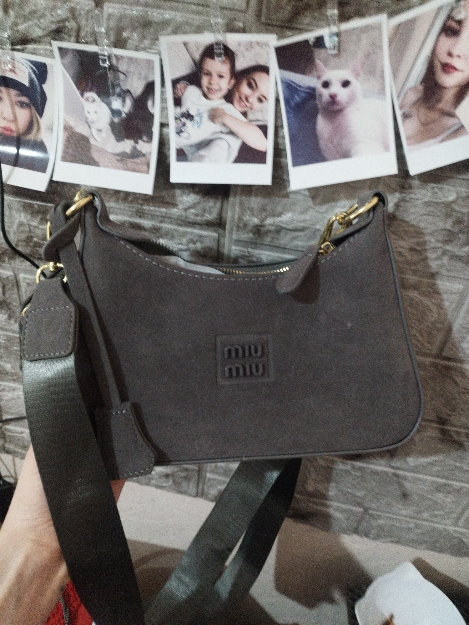 A gray Miu Miu handbag is displayed against a backdrop of a brick-like wall in what appears to be an indoor setting. The wall is decorated with photos held up by clothespins. The bag features a thick shoulder strap and gold-colored hardware.