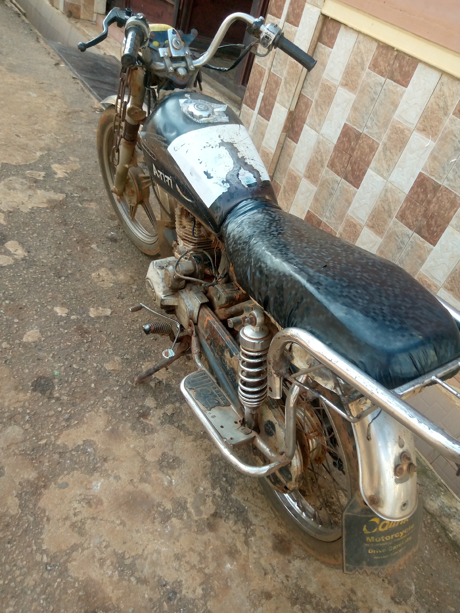 This is a medium-angle, full-body shot of a black, weathered motorcycle parked on a rough, concrete surface in Nsugpe, Nigeria. The motorcycle is the primary subject, shown from its front handlebars down to the rear tire and fender. It appears to be an older model, with visible signs of rust and wear on its metal components, including the frame, engine casing, and suspension springs. The fuel tank is black with a white, peeling sticker or paint design. The seat is black and appears to be covered with a textured material.
The motorcycle is parked next to a wall covered in a tiled pattern of beige, brown, and white. The ground beneath the motorcycle is an uneven, greyish-brown concrete with some scattered debris. The lighting suggests it is daytime, with no strong shadows indicating a specific time of day. The overall scene conveys a sense of everyday life in a location where vehicles might be used extensively and show signs of prolonged use.
There is visible text on the rear fender of the motorcycle, which reads "COMMUN Motorcycle Drive Care". The rest of the text below is too small to be legible. No people are visible in the image. The weather appears to be dry.