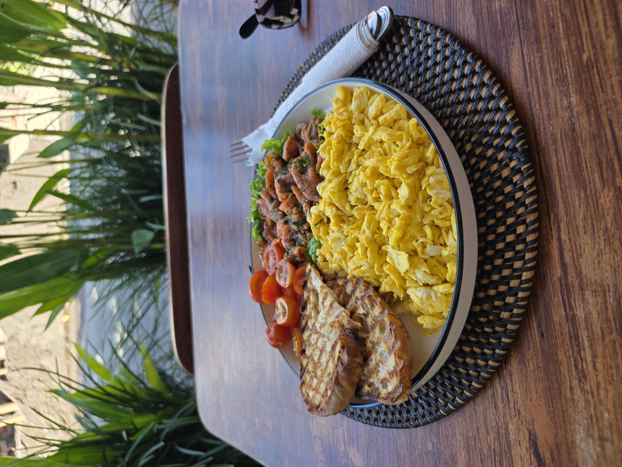 A high-angle shot captures a breakfast plate atop a wooden table. The plate contains scrambled eggs, what appears to be smoked salmon with dill, sliced tomatoes, and two pieces of grilled bread. A fork and rolled napkin are also on the table. The plate rests on a woven placemat. In the background, green foliage is visible, suggesting an outdoor or garden setting. The photograph was taken in North Kuta, Indonesia.
