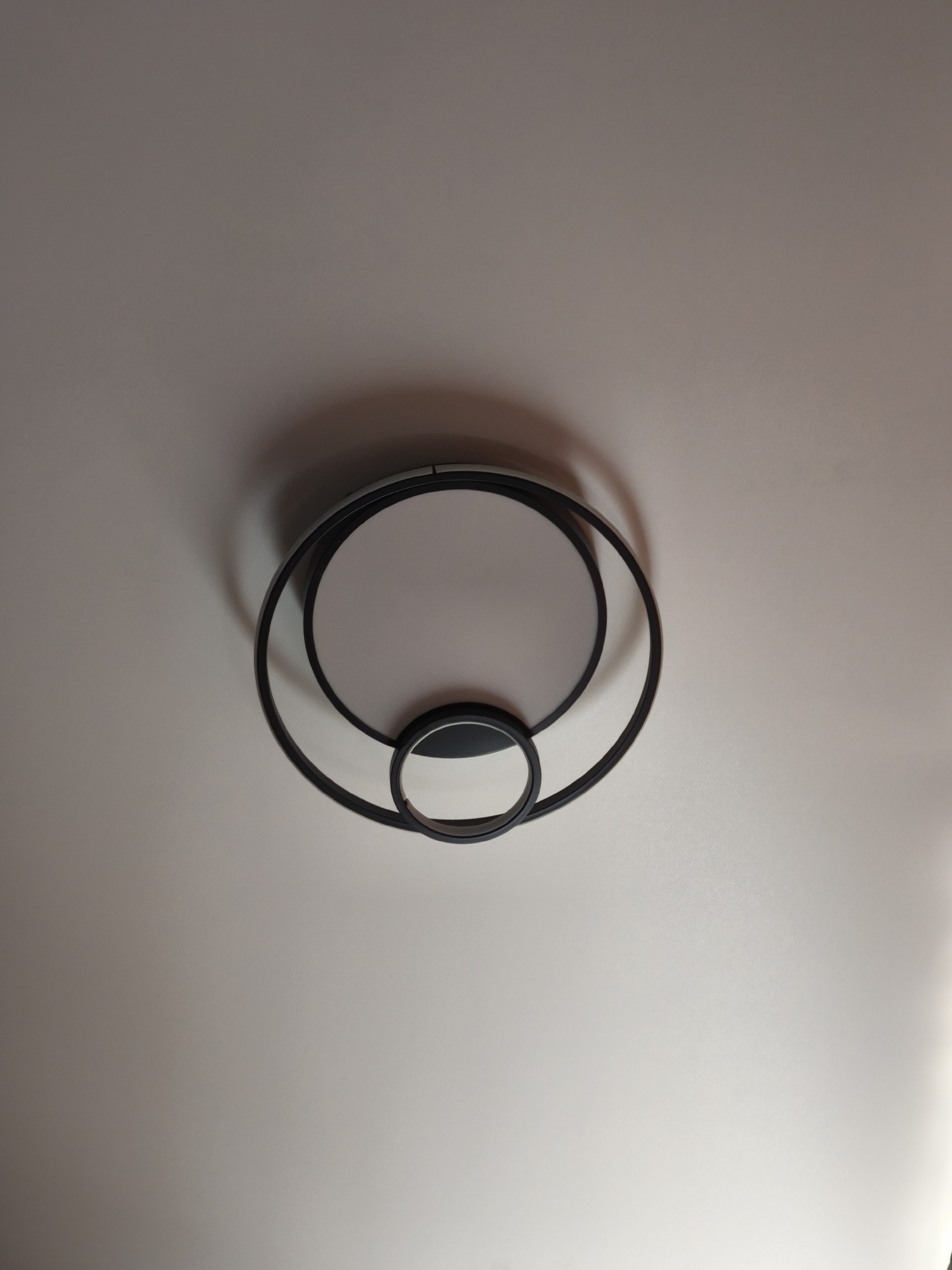 The image shows a ceiling-mounted light fixture. The fixture has two circular light panels surrounded by black metal rings. The larger light panel is centered in the fixture, and a smaller light panel is positioned below it, also encircled by a black ring. The background is a plain, white ceiling with subtle color variations due to lighting. There is no indication of the location being Moscow, Russia or any other specific city. No people, actions, or interactions are visible.