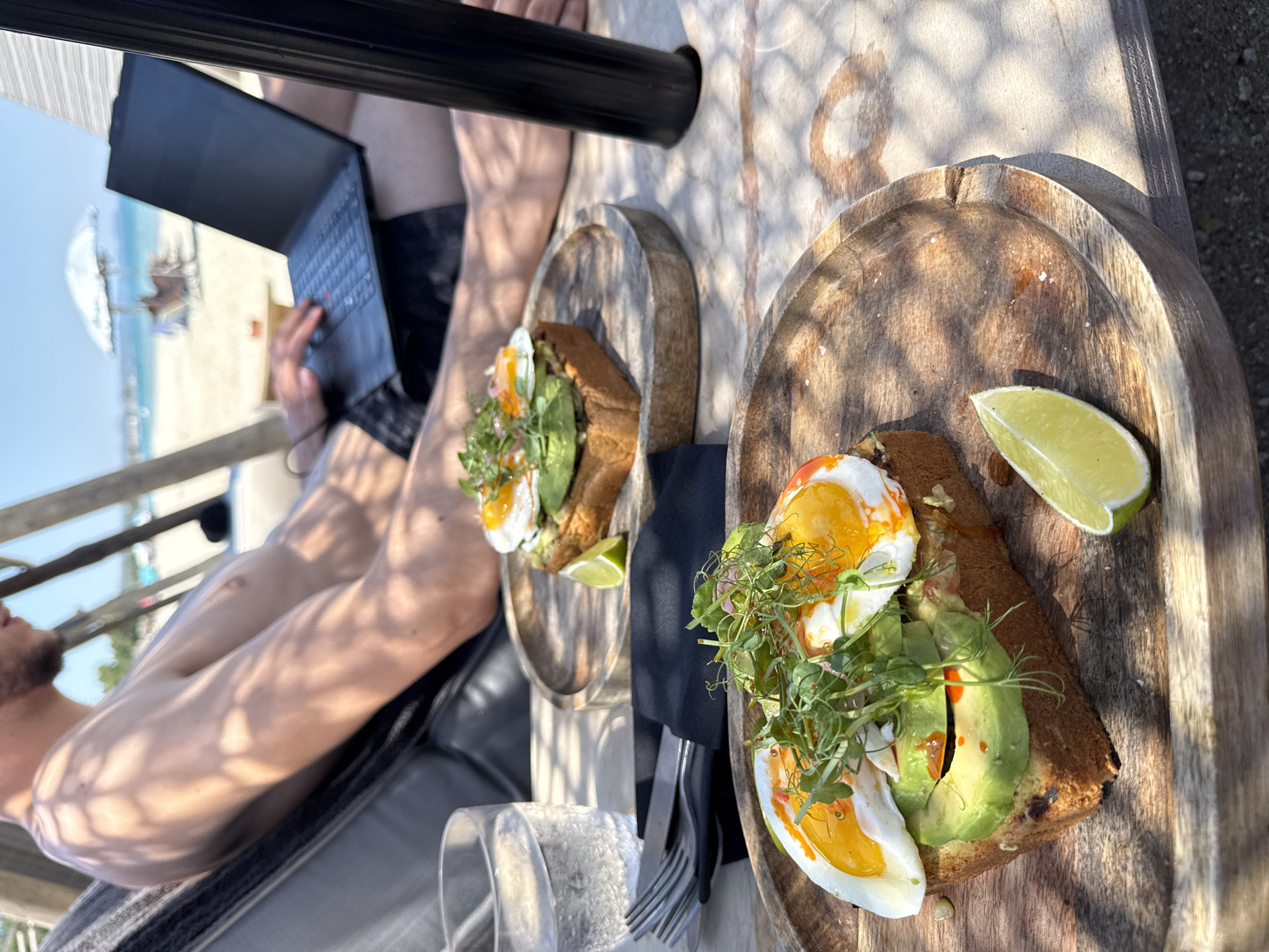 A man is reclining on a chair outdoors, looking at a laptop. In the foreground, two plates of avocado toast with eggs and microgreens are on a wooden table. A glass of water and cutlery are also on the table. The background shows a beach with an umbrella and chairs. The overall setting appears to be a beachside cafe or restaurant in Pallini Municipal Unit, Greece.
