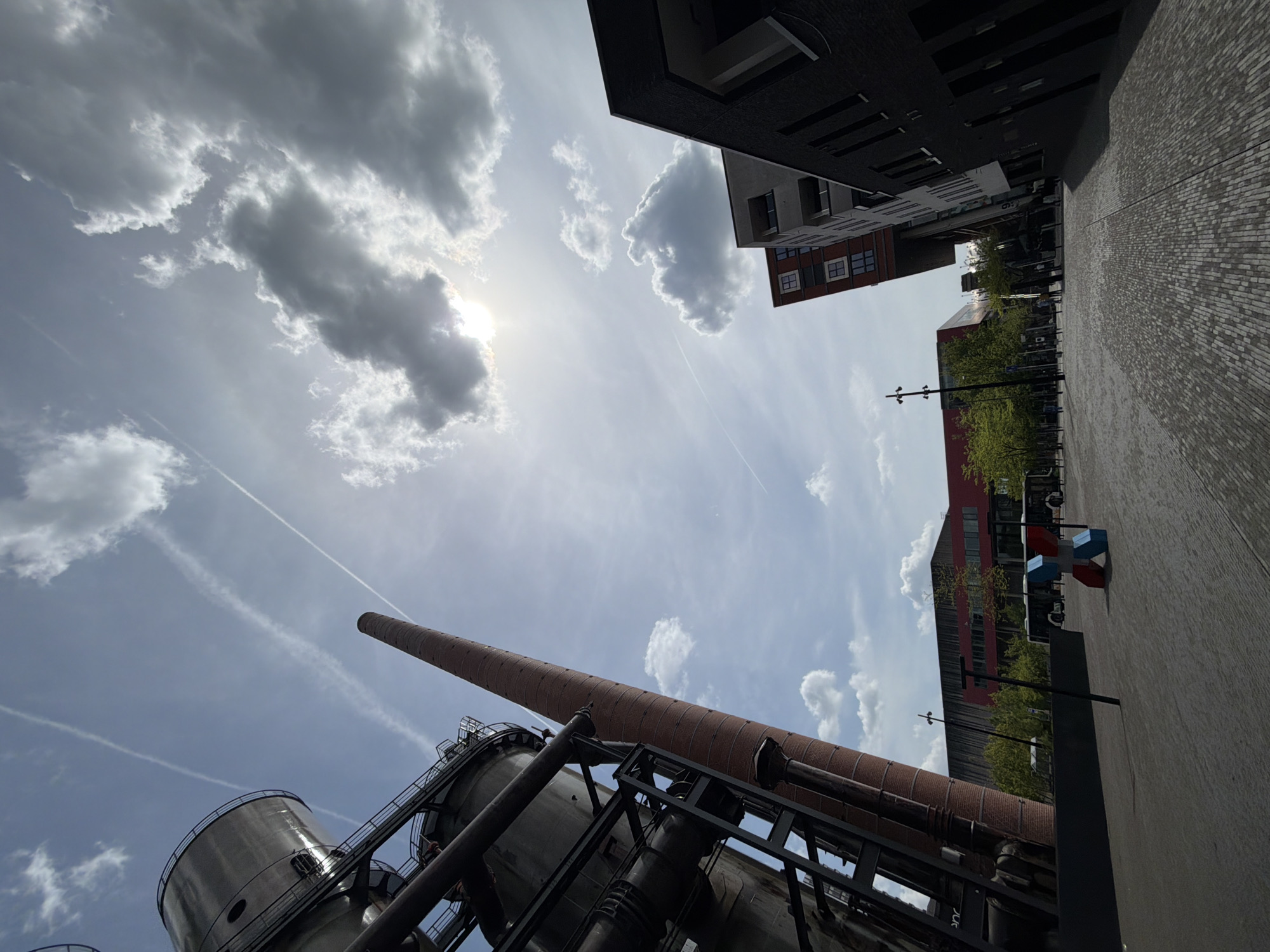 The image is a low-angle shot of the industrial heritage site in Esch-sur-Alzette, Luxembourg. The composition is upward, showcasing a towering brick smokestack and metallic industrial structures against a partly cloudy sky with contrails. Modern buildings frame the scene in the upper right and upper left. The foreground features the industrial architecture, while the background includes portions of modern buildings, trees, and the pedestrian area of the city. No people are visible.
