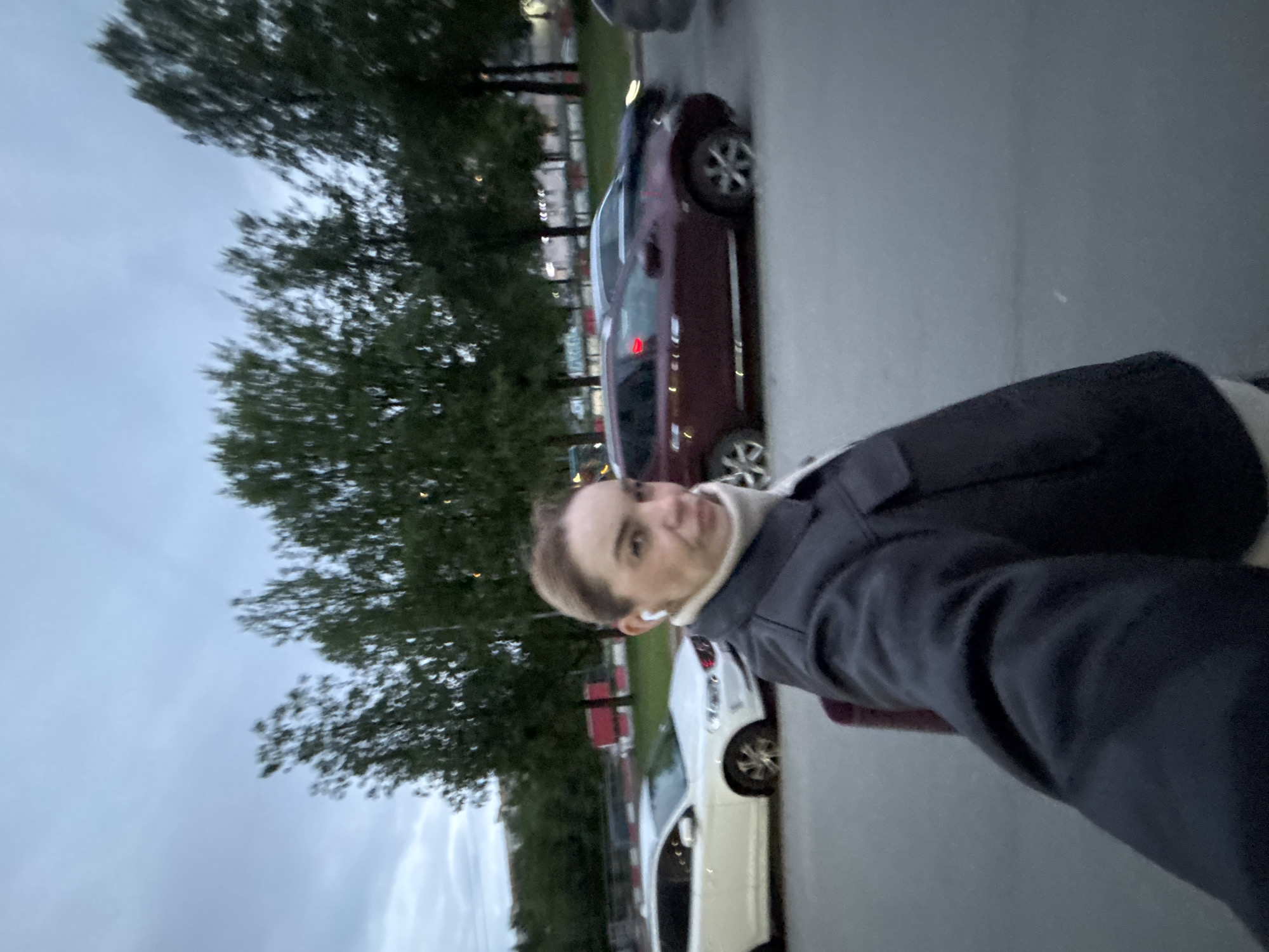 A woman takes a selfie in an urban environment near parked cars. The sky is overcast. She wears a dark jacket and white earphones. A burgundy car and a white car are parked along the roadside. Tall trees are visible in the background.