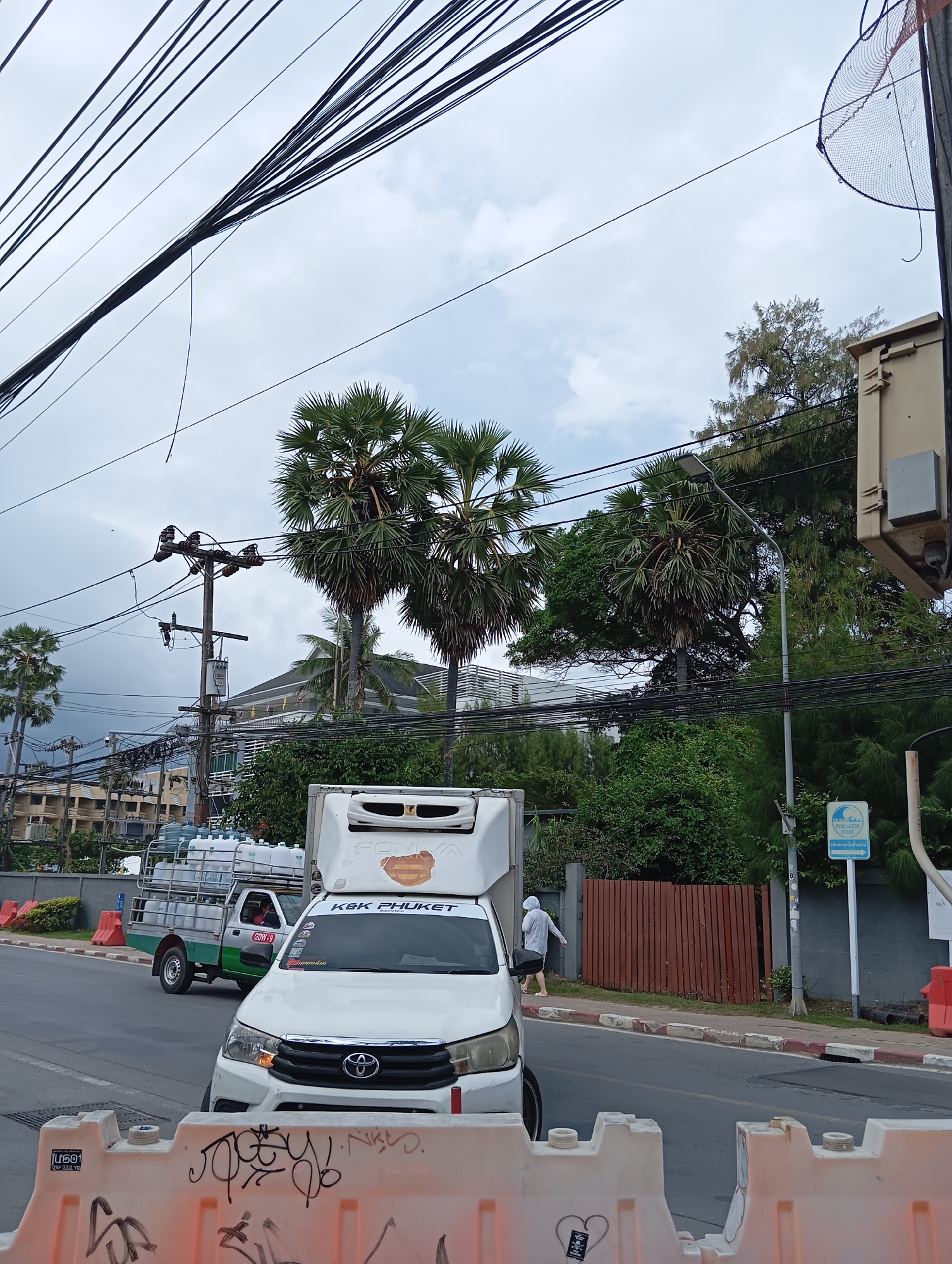 A street scene in an urban environment under an overcast sky, with numerous power lines strung across the upper portion of the image. In the foreground, a white pickup truck with a refrigerated cargo box is parked on the right side of the road, with "KBK PHUKET SERVICE" visible on its windshield. To the left, a smaller green and white pickup truck is carrying several large water jugs. A person wearing a gray hoodie and shorts walks away from the white truck on the sidewalk, which is lined with plastic barricades with graffiti. Palm trees and other greenery are visible in the background, along with buildings and utility poles.