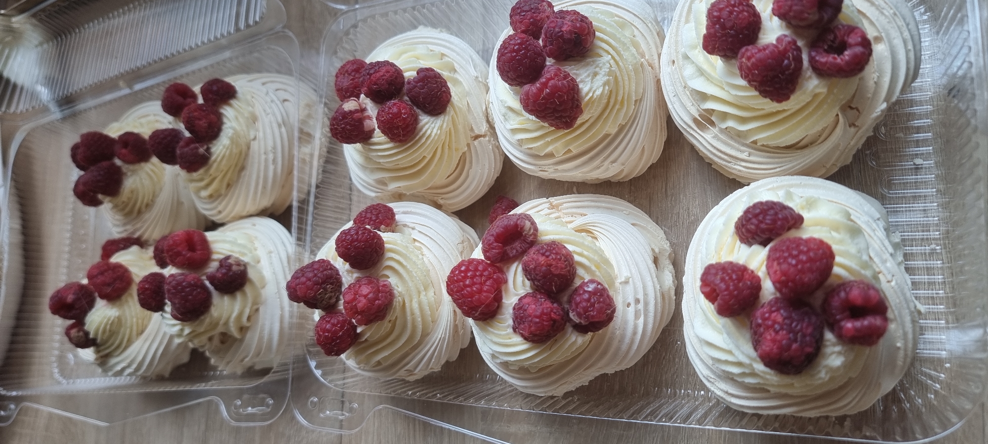 A group of pavlova desserts topped with raspberries are displayed in clear plastic containers on a wooden surface. The desserts are arranged in three rows, with four visible in the bottom row, three in the middle row, and two in the top row. Each pavlova is a swirl of meringue with a dollop of cream and several fresh raspberries on top. The lighting is natural and comes from above. There are no people or animals present in the image. The setting appears to be a kitchen or food preparation area.
