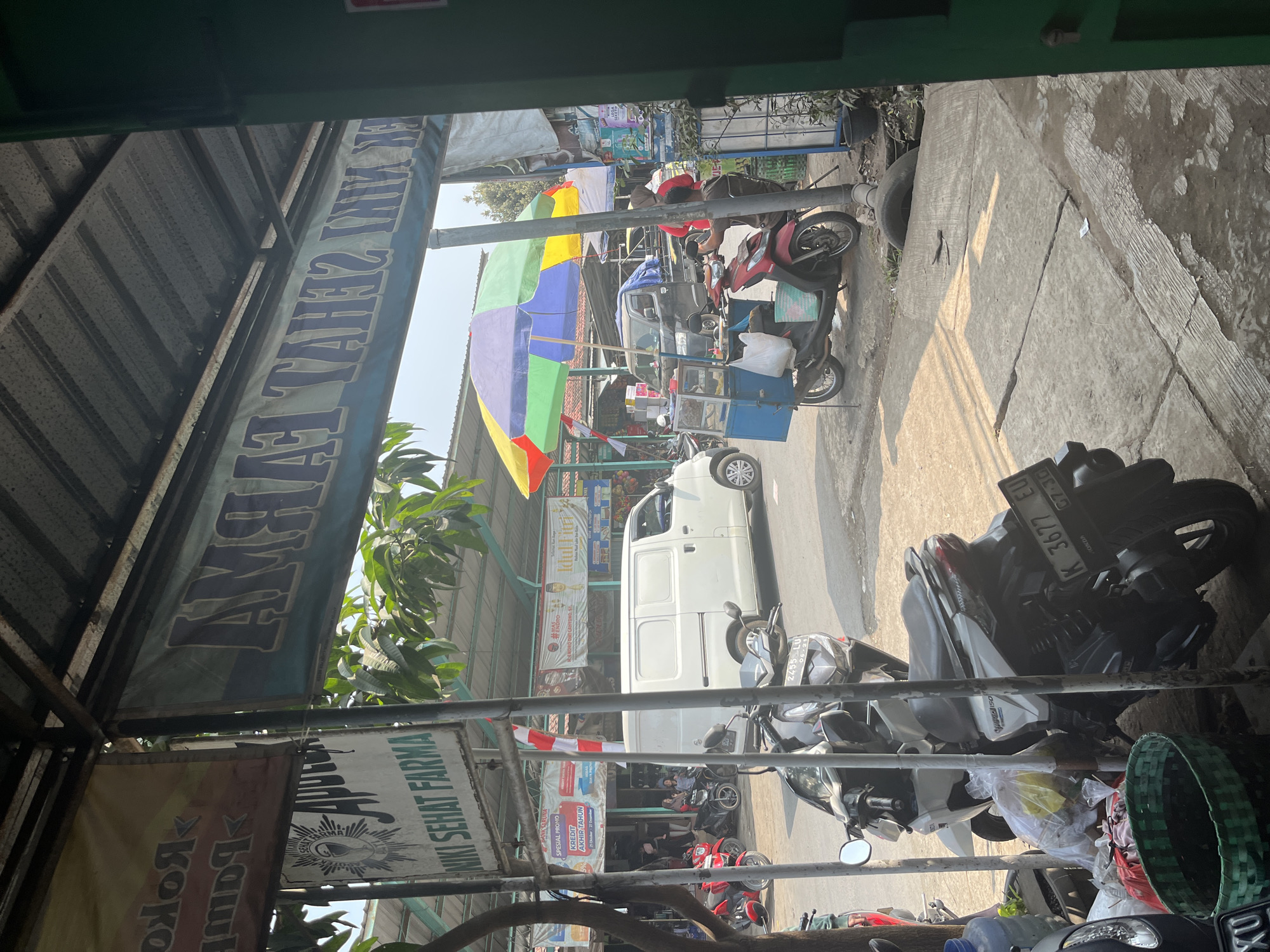 The image depicts a street scene in Gembong, Indonesia, likely a market or commercial area. In the foreground, several parked motorcycles are visible, including a white and black one with license plate K 3677 EU. To the right, a green woven basket rests near some plastic bags and trash. A large banner with blue lettering, partially obscured, hangs overhead, with Indonesian text that reads "NIKI SEHAT FARMA" and "AMHAT TAHAT SEHAT INIK".

Further down the street, a white van is parked, and a food cart with a colorful umbrella is set up. A person wearing a red hijab is standing near the food cart, and another individual in a brown shirt appears to be interacting with it. Red motorcycles are also parked on the left side of the street. Several other banners and signs are visible in the background, indicating shops or stalls. The environment appears to be outdoors, with a paved road and concrete walkways, under a bright sky. The overall scene suggests a busy daytime commercial setting.