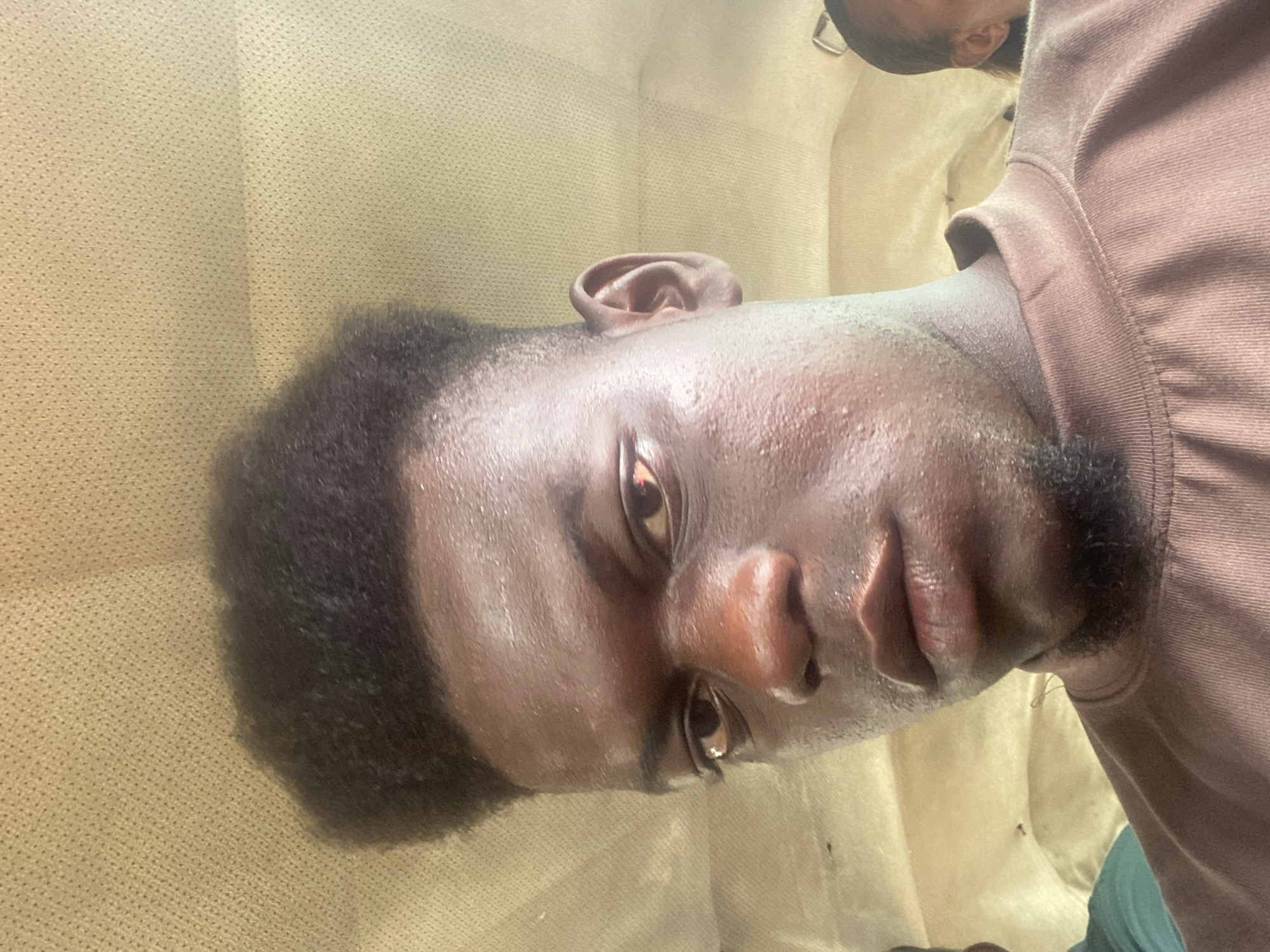 A close-up photograph shows a dark-skinned man with short, dark hair and a beard stubble. He is wearing a light brown shirt. The background appears to be a tan, textured fabric, possibly a couch. The location is unknown.