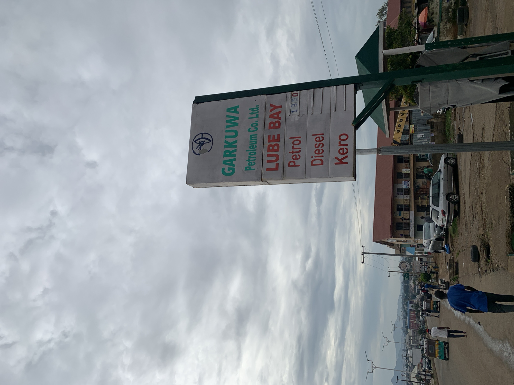 This is a street-level, eye-level, medium shot captured on a cloudy day in the city of Rafin Par, Nigeria. The primary subject is a gas station sign on the right side of the image. The sign is white with green and red text. The top section reads "GARKUWA Petroleum Co. Ltd.". Below that, it says "LUBE BAY". The next two lines, "Petrol" and "Diesel", are in red text. Next to "Petrol" is a price of "1350" in smaller text. The bottom line reads "Kero" in red text.
In the background, a street is visible with several vehicles, including cars, a tuk-tuk, and a truck. People are walking along the street and sidewalks. Buildings with red-tiled roofs line the street. The sky is overcast with a layer of gray clouds. The overall environment suggests an active urban setting.