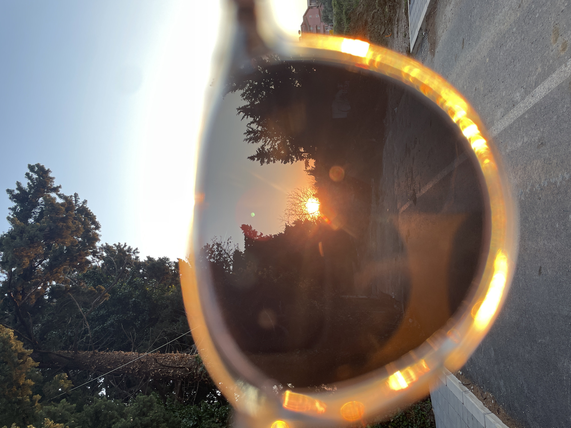The image shows a pair of sunglasses held up against a sunset view, likely in the city of Bar, Montenegro. The lens reflects the silhouette of trees with the sun setting behind them. Outside the sunglasses' reflection, there are more trees and a clear sky to the left, suggesting a late afternoon or early evening setting. To the right of the sunglasses' reflection, asphalt and a low concrete wall are visible. The city of Bar is assumed based on geographical cues such as vegetation, buildings, and the sun's position and elevation.