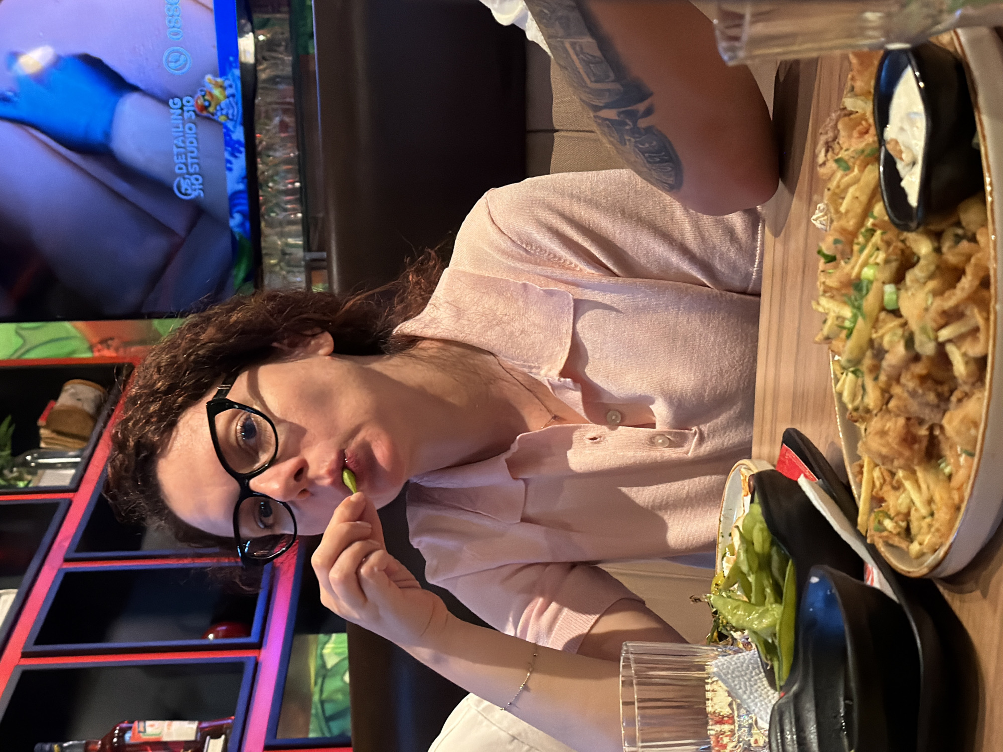 A woman with curly brown hair and black-rimmed glasses is seated at a table, eating green beans. She is wearing a light pink collared shirt. In front of her, on the table, are several bowls of food, including edamame and fried calamari. A glass of water is also visible. In the background, a shelf unit with shelves lit by red and blue lights is visible. The woman's left arm is extended towards her face, holding a green bean to her mouth. The scene appears to be in a restaurant.