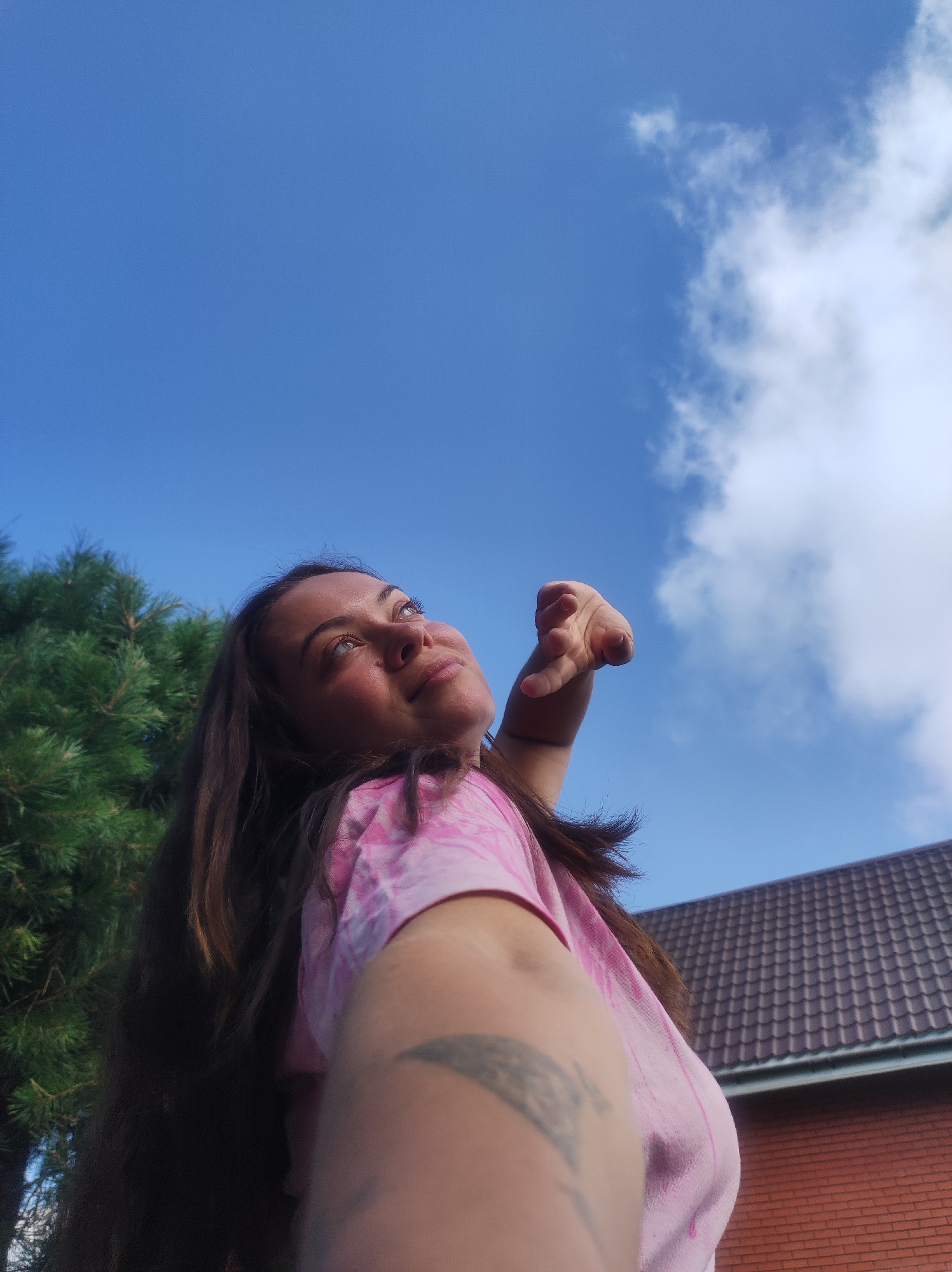 The image shows a young woman with long brown hair, wearing a pink tie-dye t-shirt, looking up at the sky. Her right hand is raised and extended towards the sky, with her fingers slightly curled. She has a tattoo on her left forearm. The background consists of a bright blue sky with scattered white clouds, green pine trees to the left, and a brown-tiled roof of a building to the right. The perspective suggests the photo was taken from a low angle, looking upwards. The scene appears to be outdoors, possibly in a residential area of the city of Fokino, Russia, given the presence of a building's roof and mature trees.