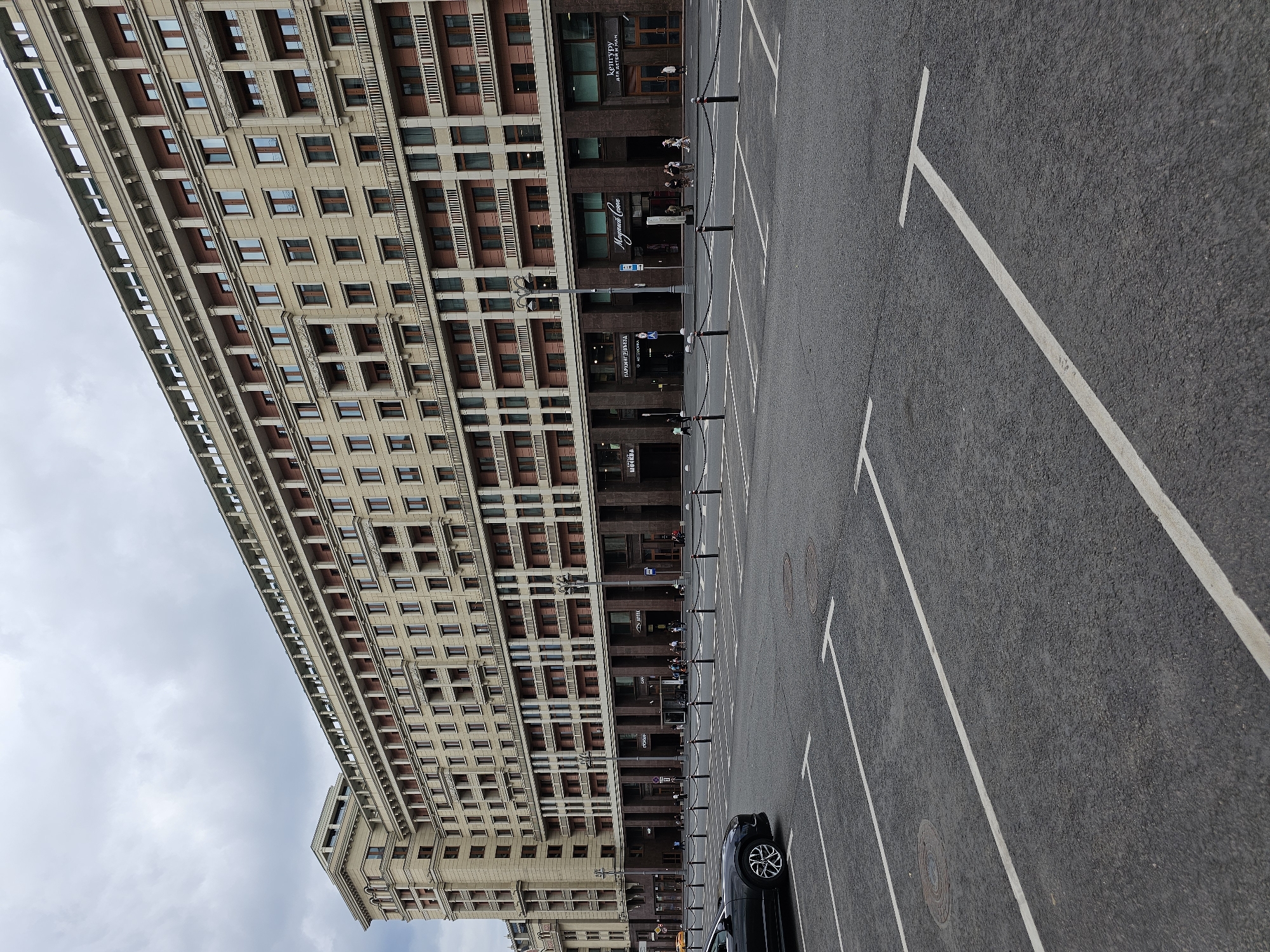 The image shows a wide, empty parking lot in front of a large, ornate, light-colored building. The building appears to be a hotel or a large apartment complex, with many windows and balconies. The architecture is grand and classical, with columns and decorative cornices. The sky is overcast and gray. A black car is parked on the left side of the parking lot. White lines mark out parking spaces. In the background, several people are visible walking on the sidewalk in front of the building. The building has storefronts on the ground level, including one with the sign "МАРГРИТ СОКОЛ" and another "КЕНТУРУ ДЛЯ ДЕТЕЙ И МАМ". Another sign visible is "АПТЕКА" (Pharmacy). The scene is located in Moscow, Russia, as indicated by the Cyrillic signage.