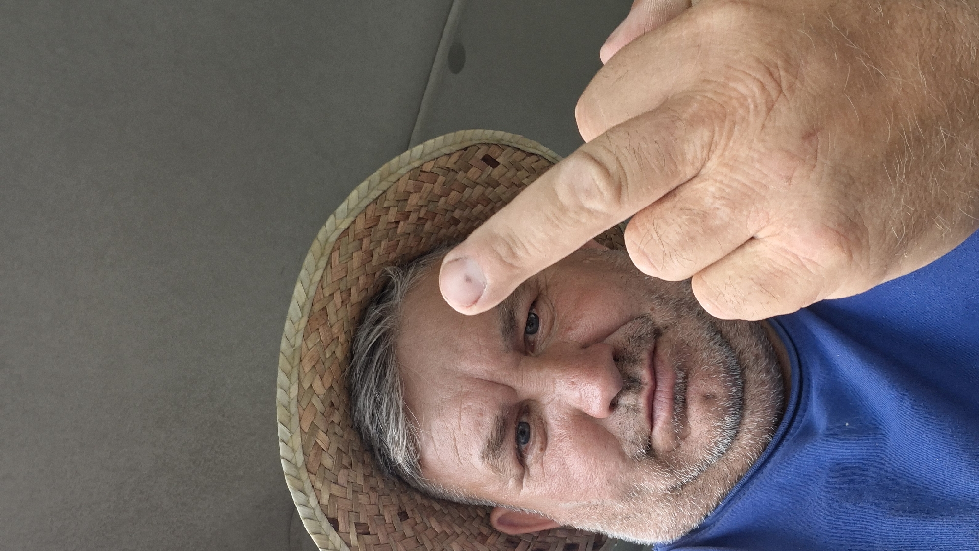A man wearing a straw hat and a blue t-shirt is captured in a close-up, selfie-style photograph. He has graying hair and a beard, and is looking directly at the camera with a neutral expression. His right hand is raised, and his middle finger is extended towards the viewer. The background appears to be the interior of a vehicle. The image does not contain enough information to determine a specific location in Hîncești, Moldova.