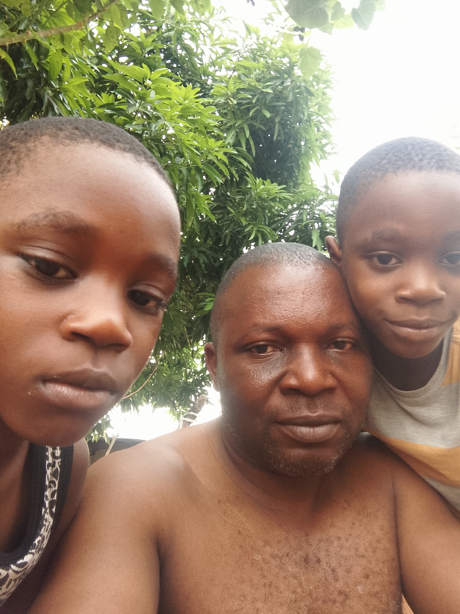 Three people are visible in the image. Two children are on the left and right sides of the image. A man stands in the center between the two children. A tree is behind the people. The location is unknown.