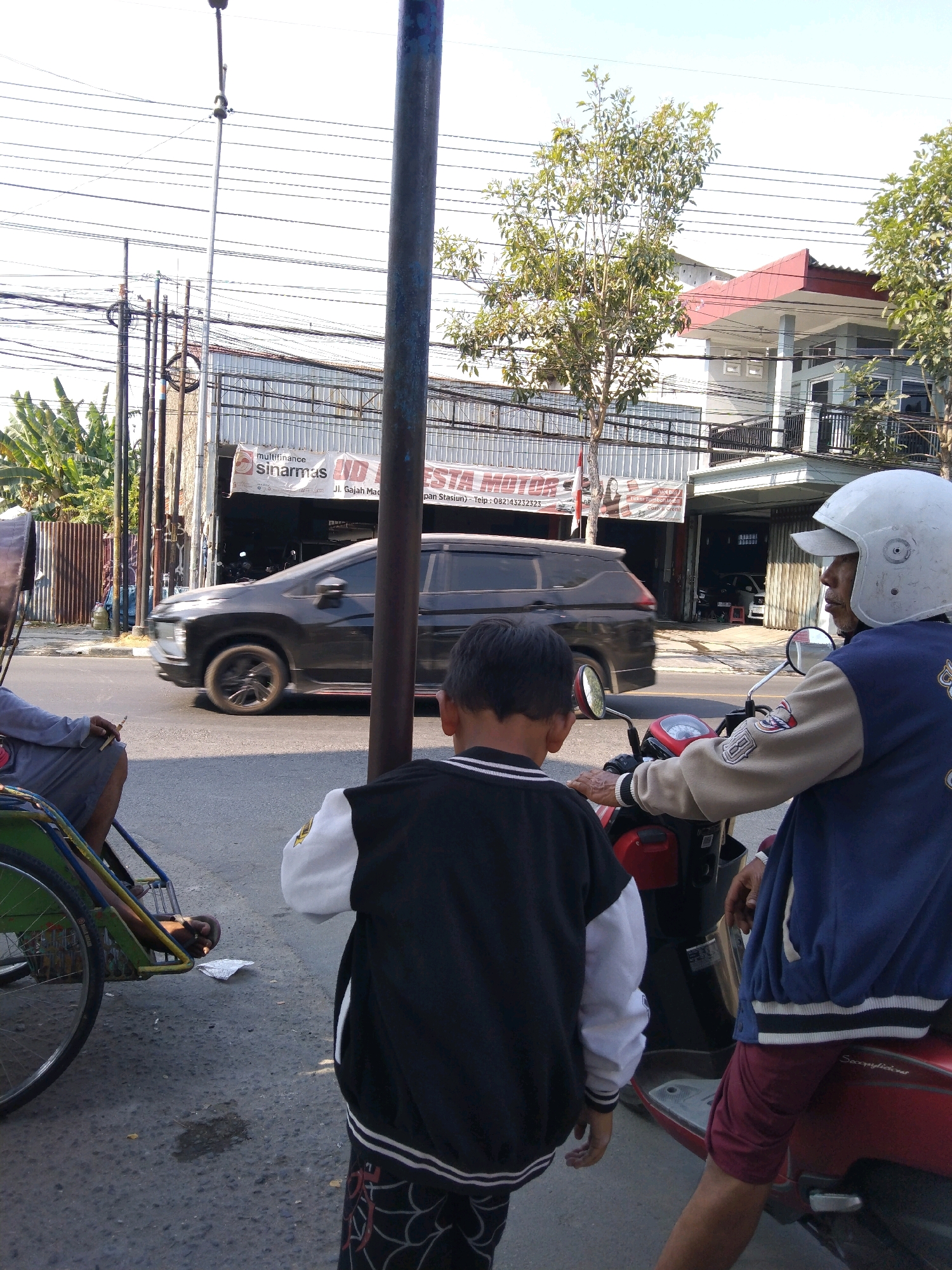 In Bojonegoro, Indonesia, a street scene unfolds on a sunny day. A dark-colored MPV drives past on the road, its wheels blurred due to motion. On the left, a man sits in a pedicab, holding a cigarette. In the foreground, a young boy stands facing away from the camera, wearing a black and white jacket. To the right, a man wearing a white helmet and a blue jacket sits on a red motorcycle, looking to the right. Behind them, a building with a red-tiled roof and a metal awning is visible, with a banner that reads "Multifinance Sinarmas". Several utility poles with tangled wires are also present. The ground is paved asphalt, with some debris and a dark stain visible.