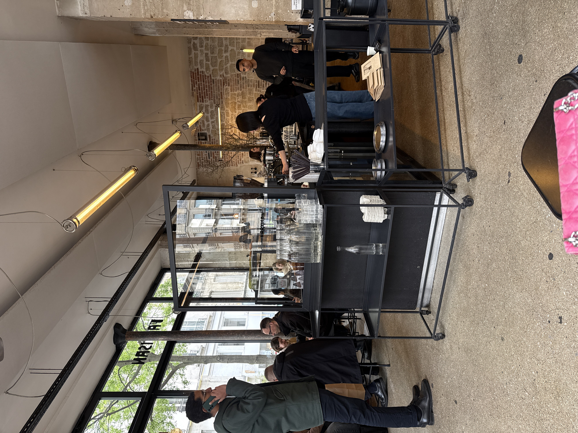 The image shows the interior of a café in Paris, France. The café has large windows that offer a view of the street outside. Inside, customers are seated at tables, some are talking on their phones, while others appear to be in conversation. Behind the customers, employees are working behind a counter, seemingly preparing drinks. The café's design includes exposed brick walls, modern lighting fixtures, and metal shelving displaying glassware and other items.